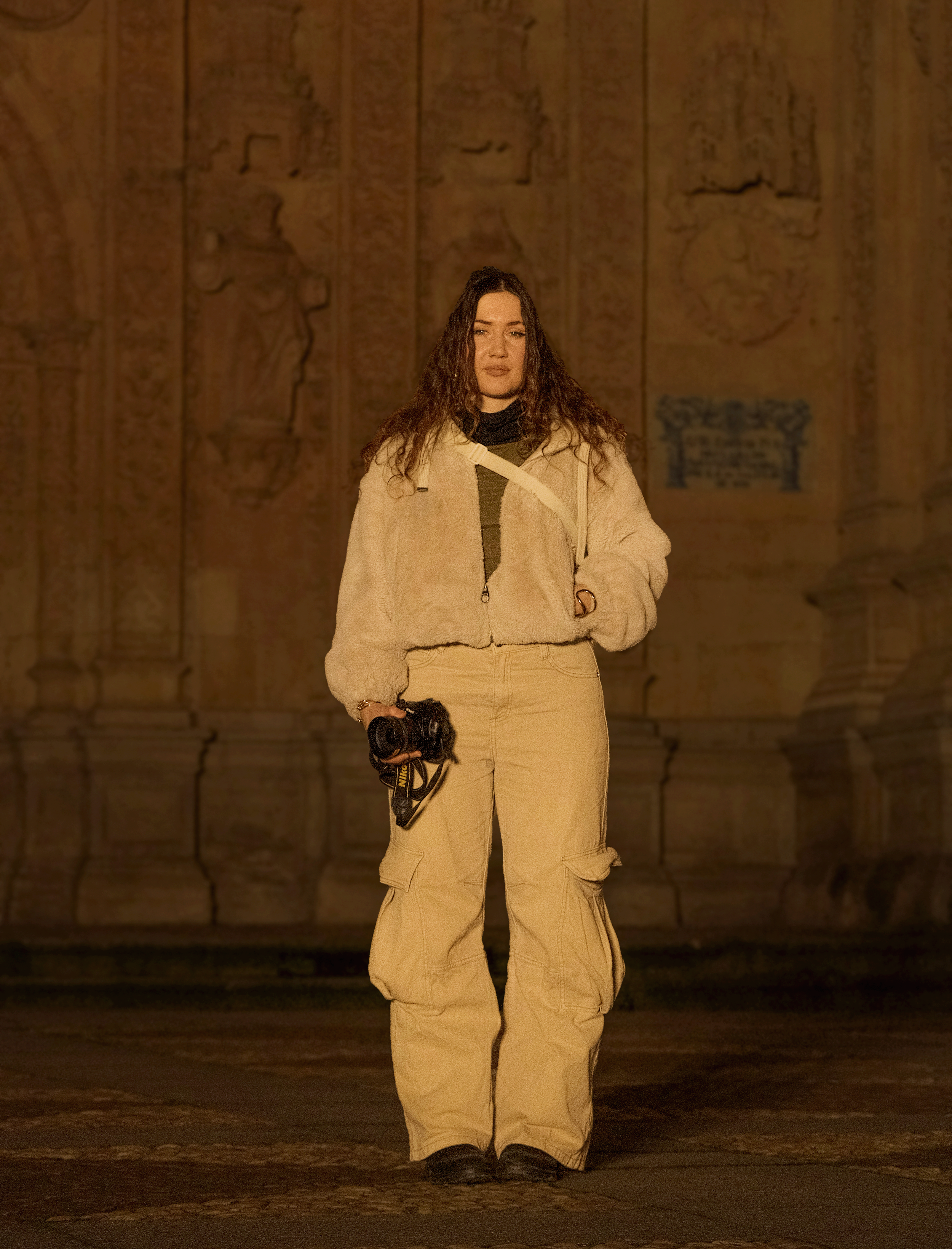 A woman standing outdoors at night in front of an ornate, illuminated stone wall. She is holding a camera in her right hand and is dressed in casual beige cargo pants and a beige fleece jacket.