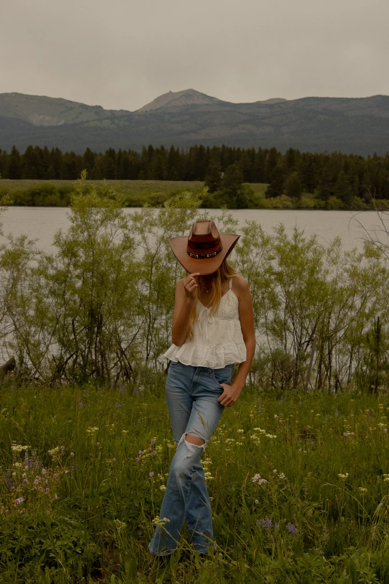 A woman standing in a field of tall grass and wildflowers, wearing a white sleeveless top, ripped jeans, and a large cowboy hat, with a lake and mountains in the background.