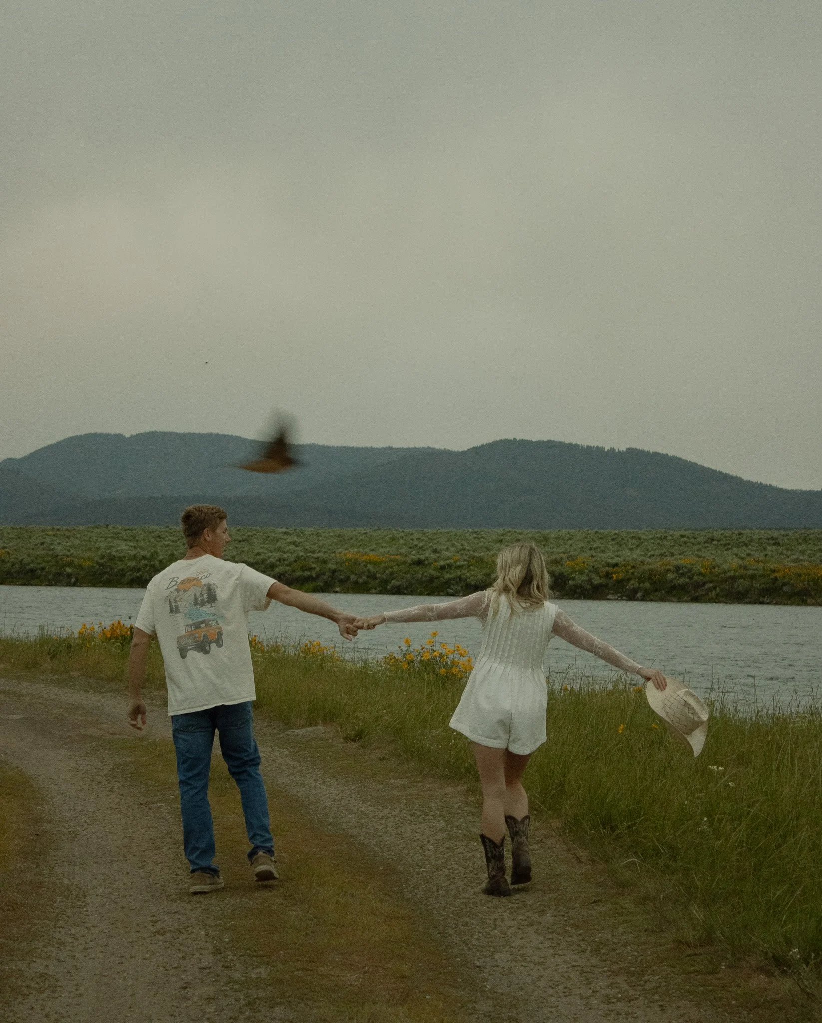 A young couple holding hands and walking along a dirt path beside a body of water, with mountains in the background and a bird flying above.