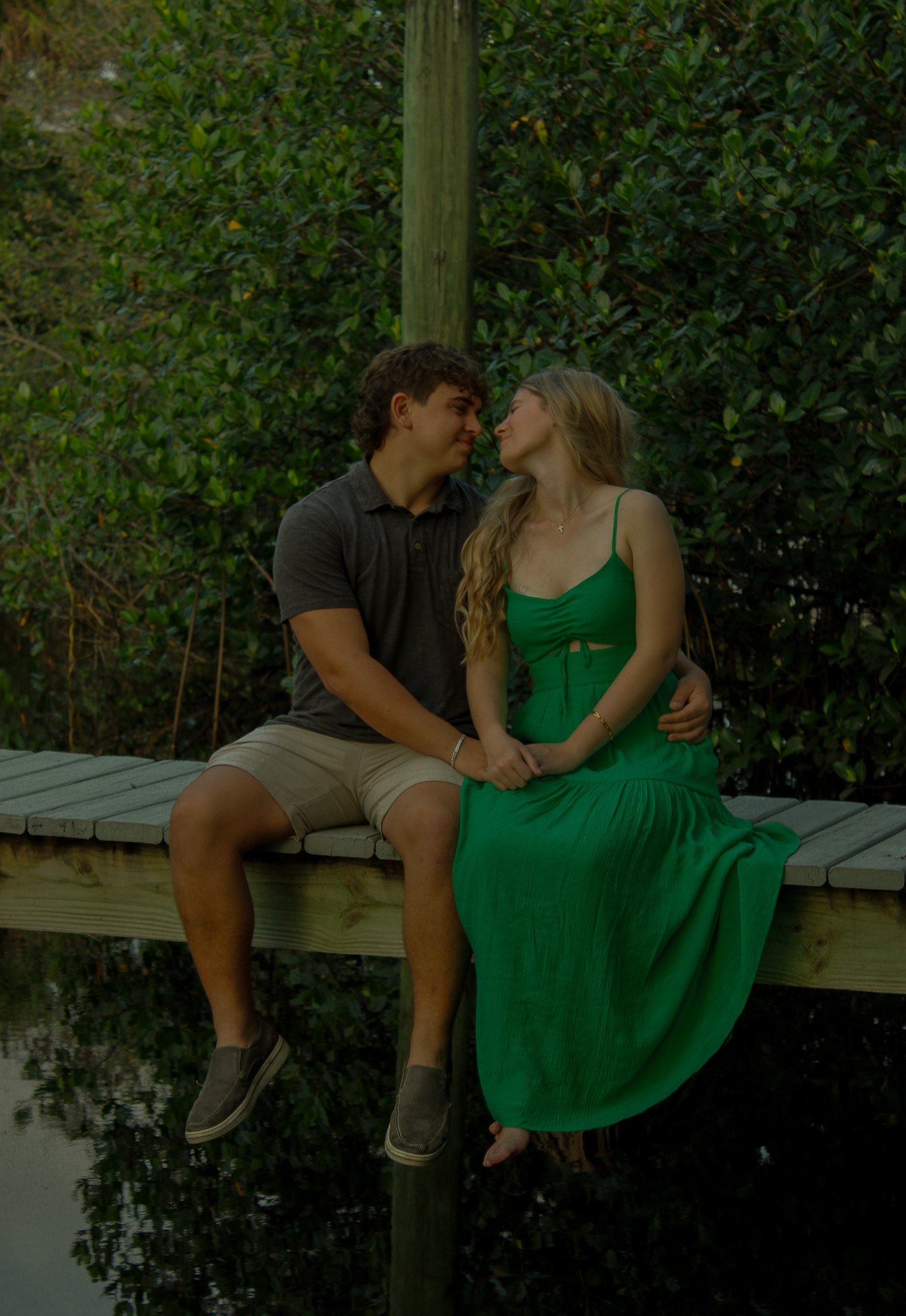 A young man and woman sit closely on a wooden dock, touching hands, with their foreheads tilted towards each other, surrounded by green foliage, suggesting an intimate moment by the water.