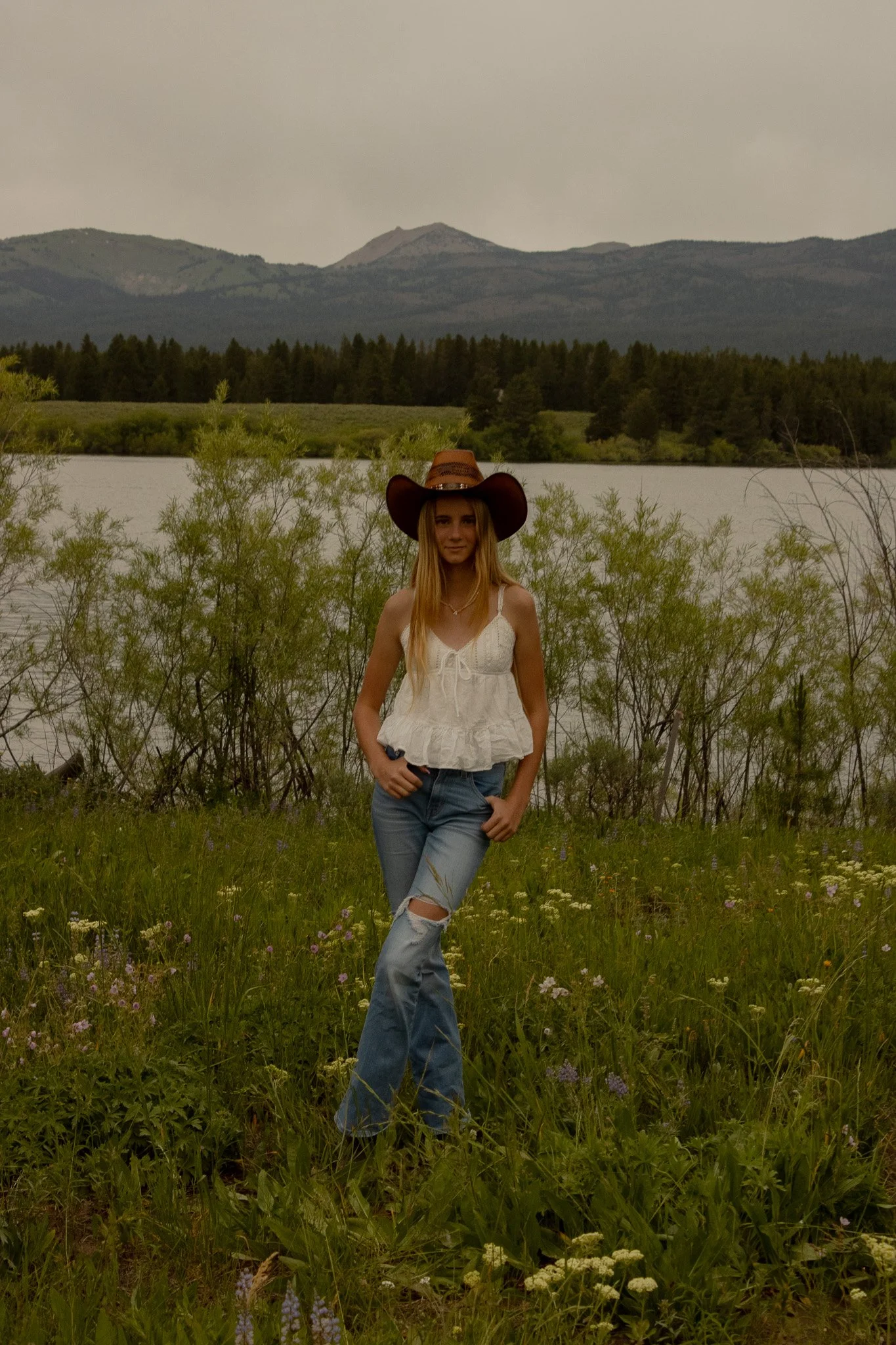 Young woman wearing a cowboy hat and white top standing in a grassy field with wildflowers, near a lake with mountains in the background.