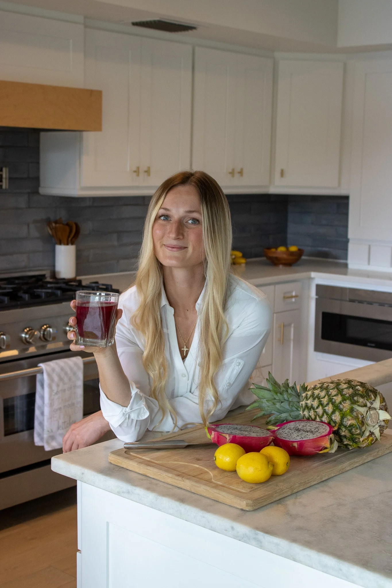 A woman with long blonde hair and light skin sitting in a kitchen, holding a glass of red juice, with a cutting board of tropical fruits including pineapple, dragon fruit, and lemons in front of her.