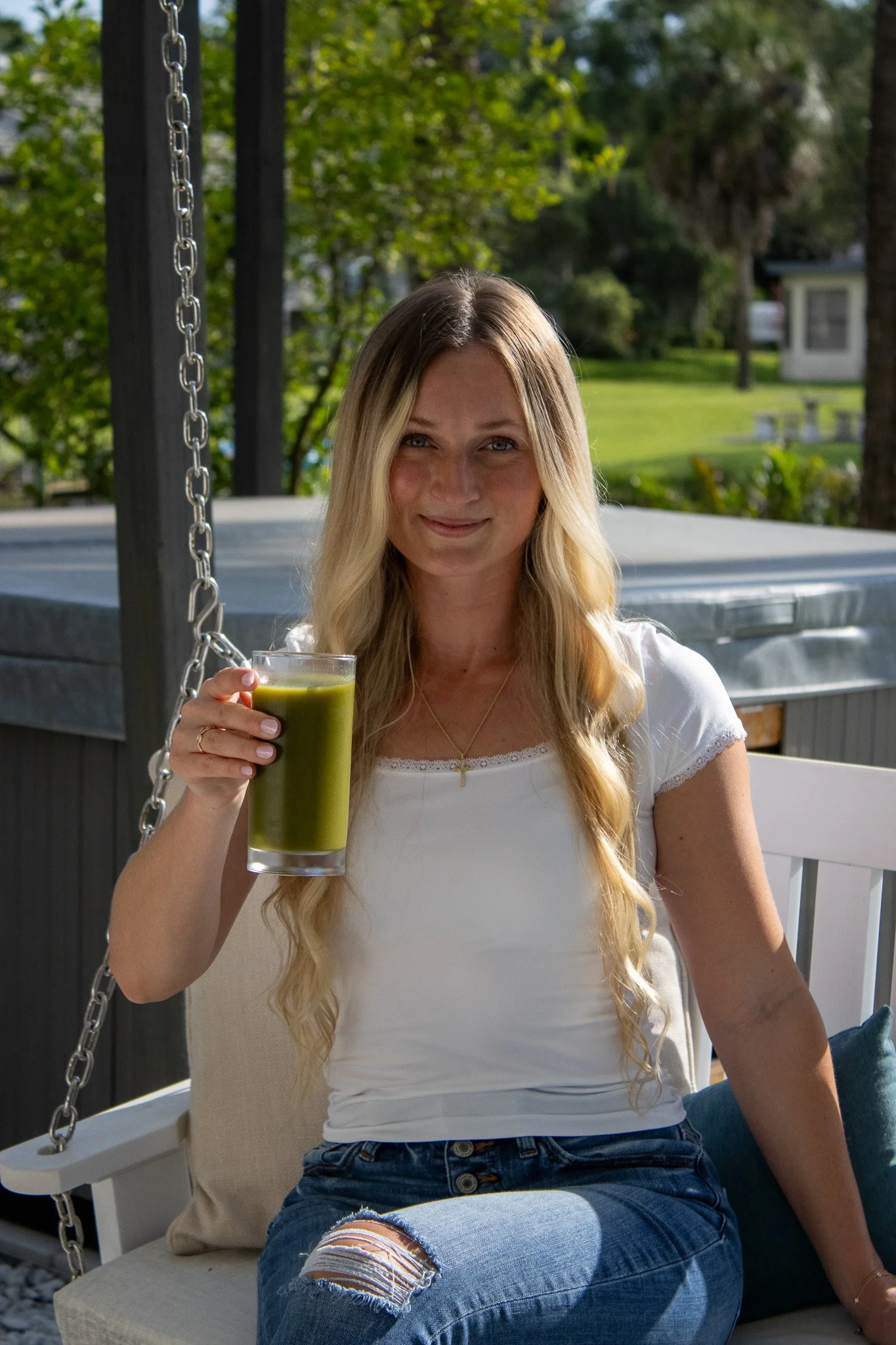 A woman sitting on a porch swing outdoors, holding a glass of green juice and smiling at the camera.