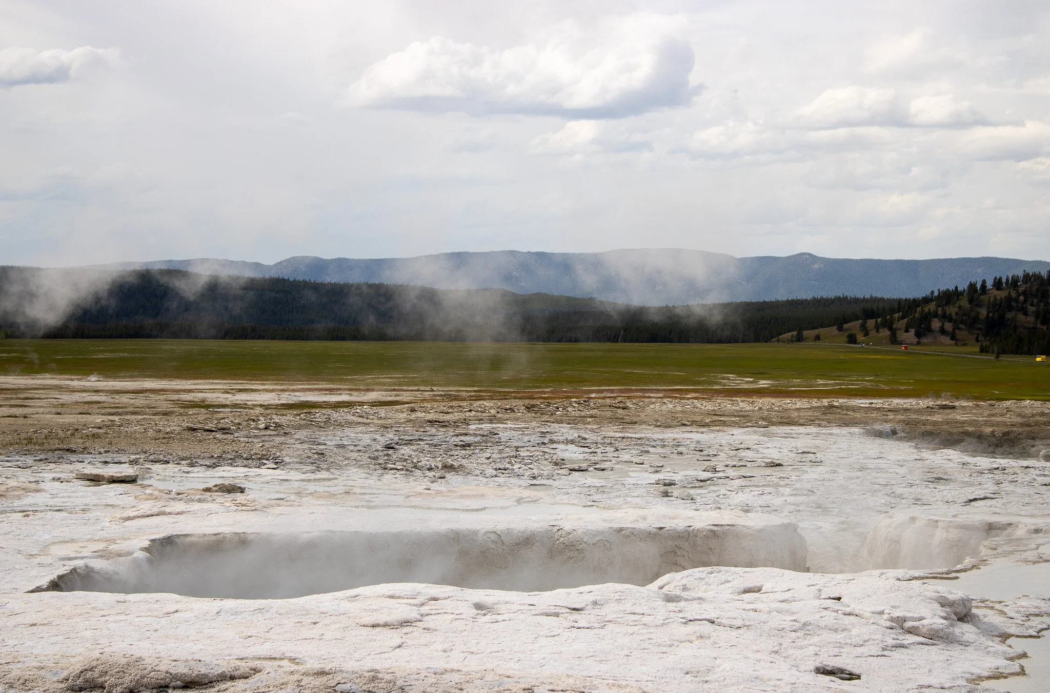 Geyser erupting in a geothermal area with steaming vents, surrounded by flat terrain, distant green hills, and cloudy sky.