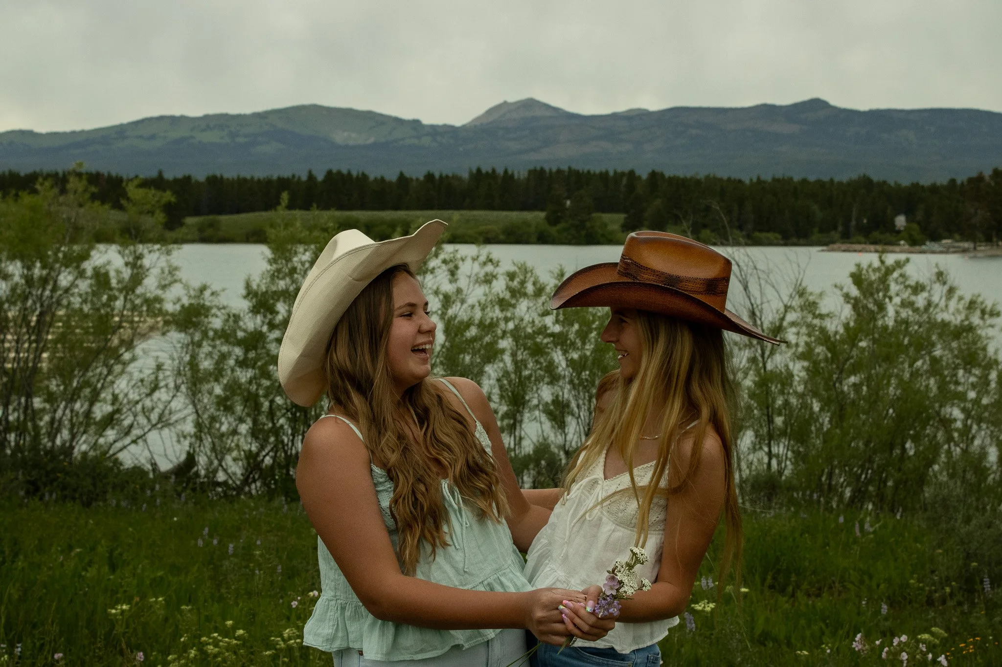 Two young women wearing cowboy hats smiling and holding flowers by a lake with mountains in the background.