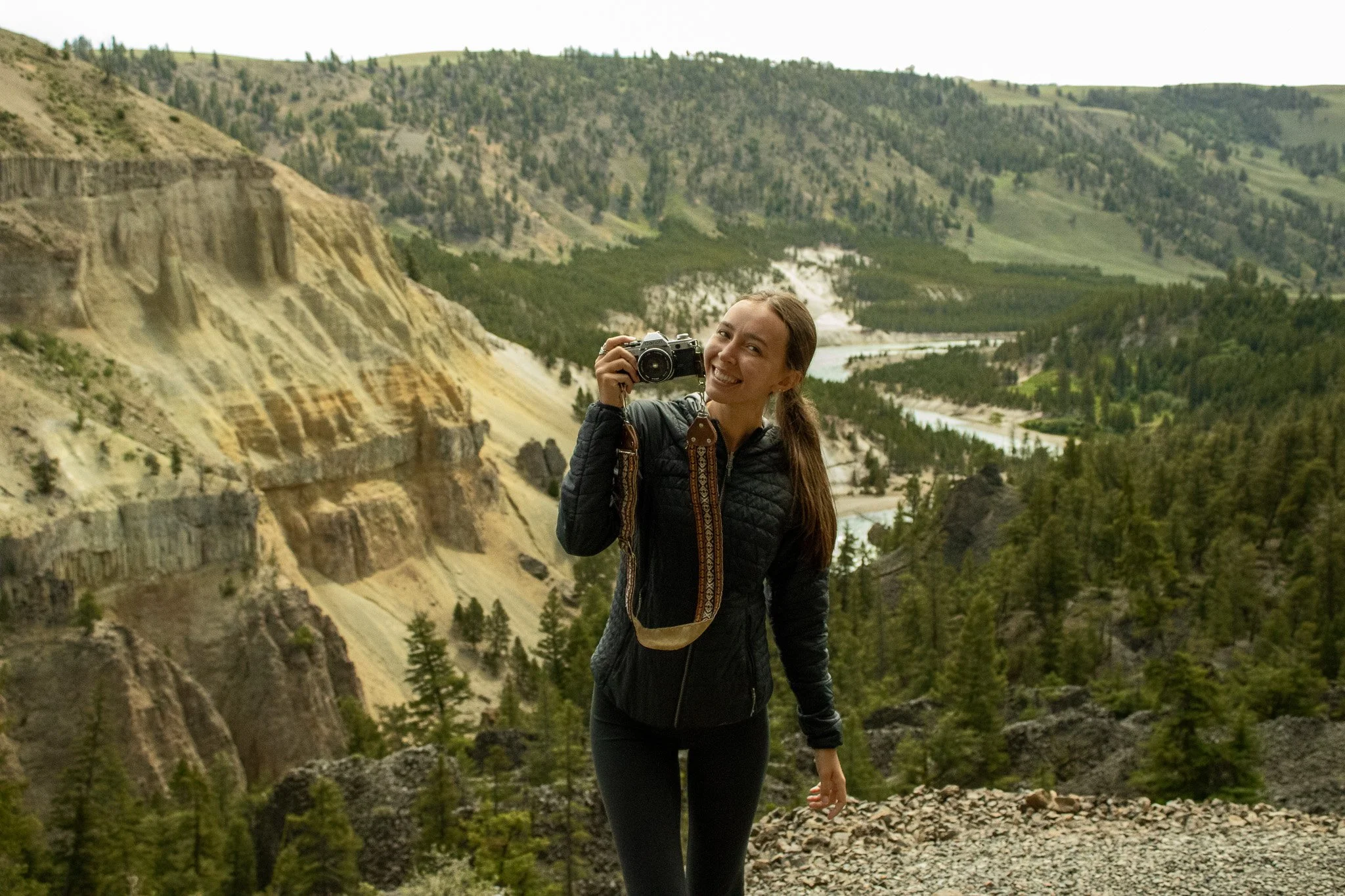 Alyssa smiling and holding a camera in a mountain landscape with forests and a river in the background.