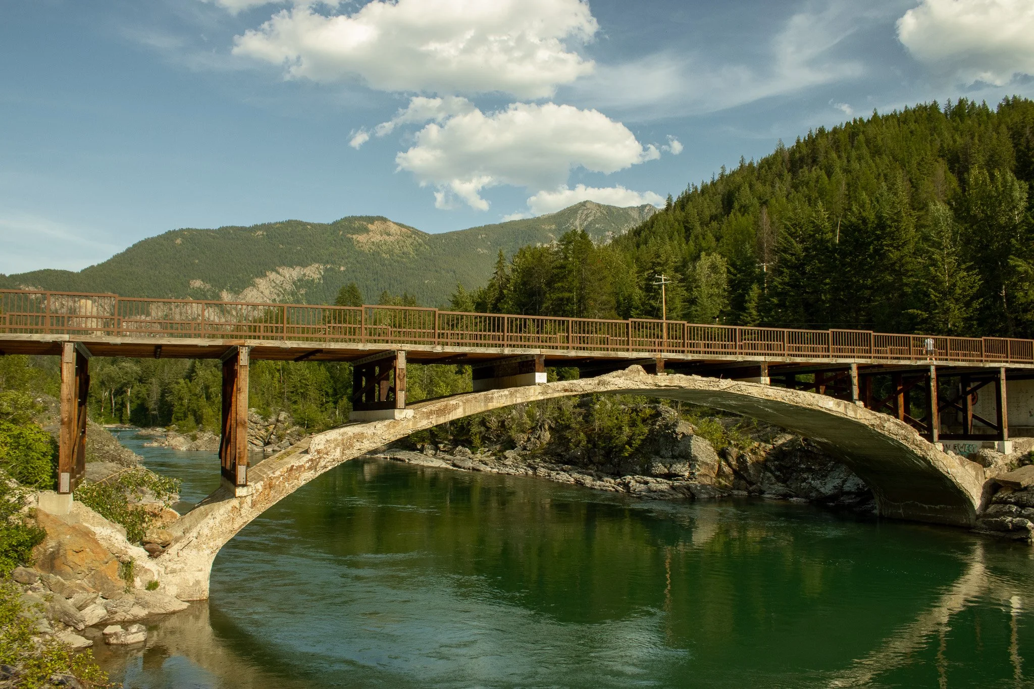 A stone bridge over a river with dense green forest and mountains in the background under a partly cloudy sky.