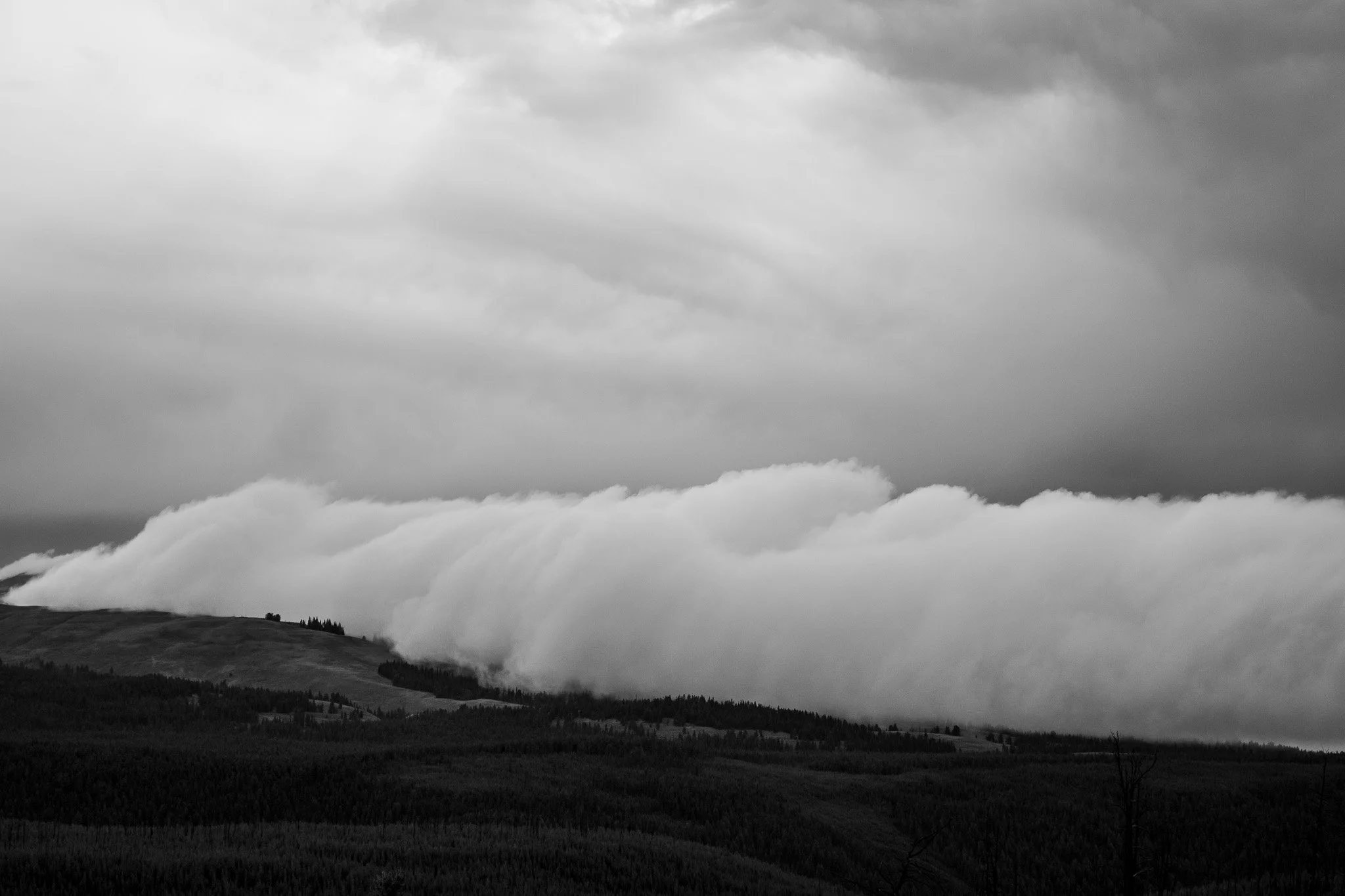 A black and white photograph of rolling hills beneath a dramatic sky with thick, layered clouds.