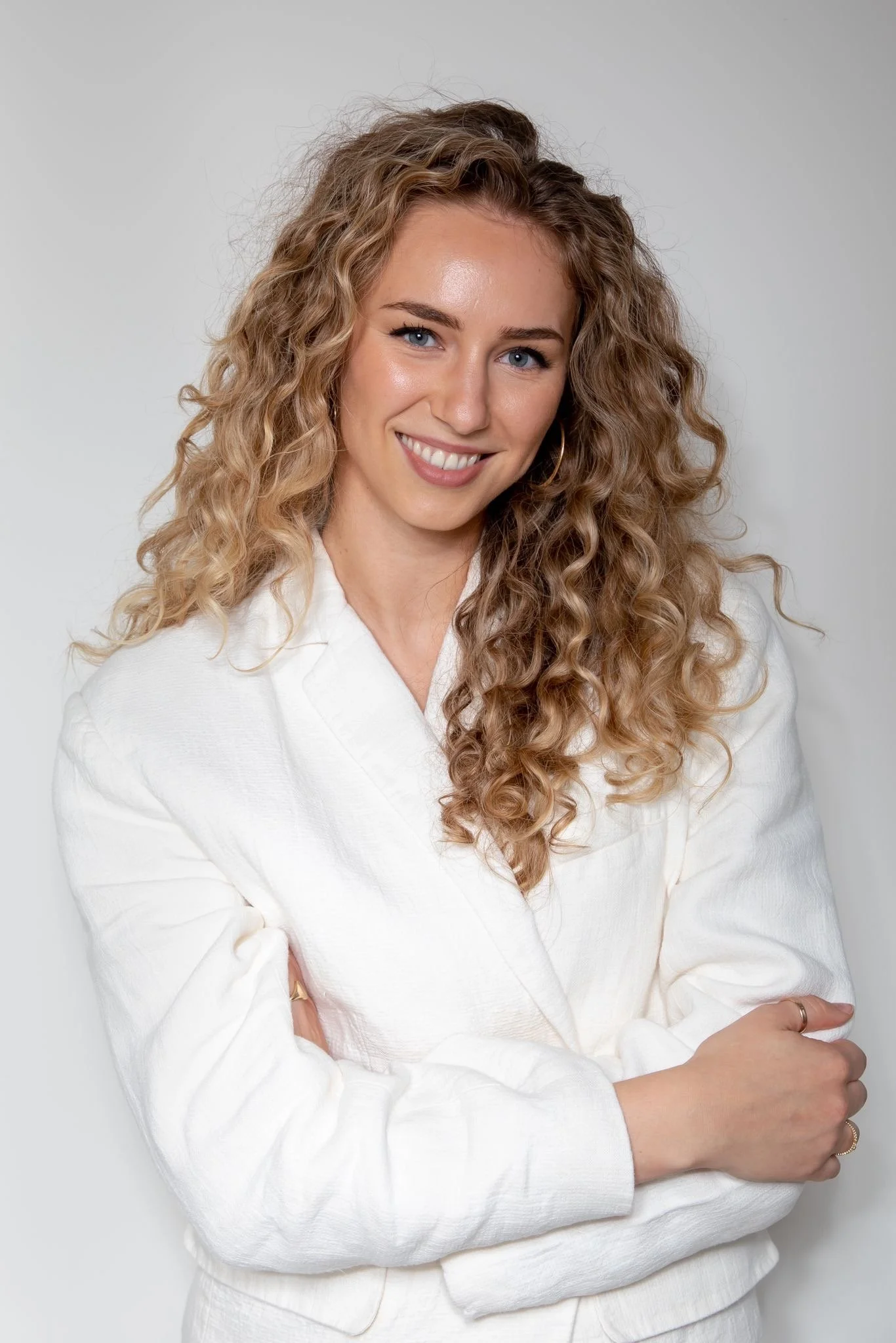 Portrait of a woman with long, curly blonde hair, wearing a white blazer, smiling, and standing against a plain light gray background.