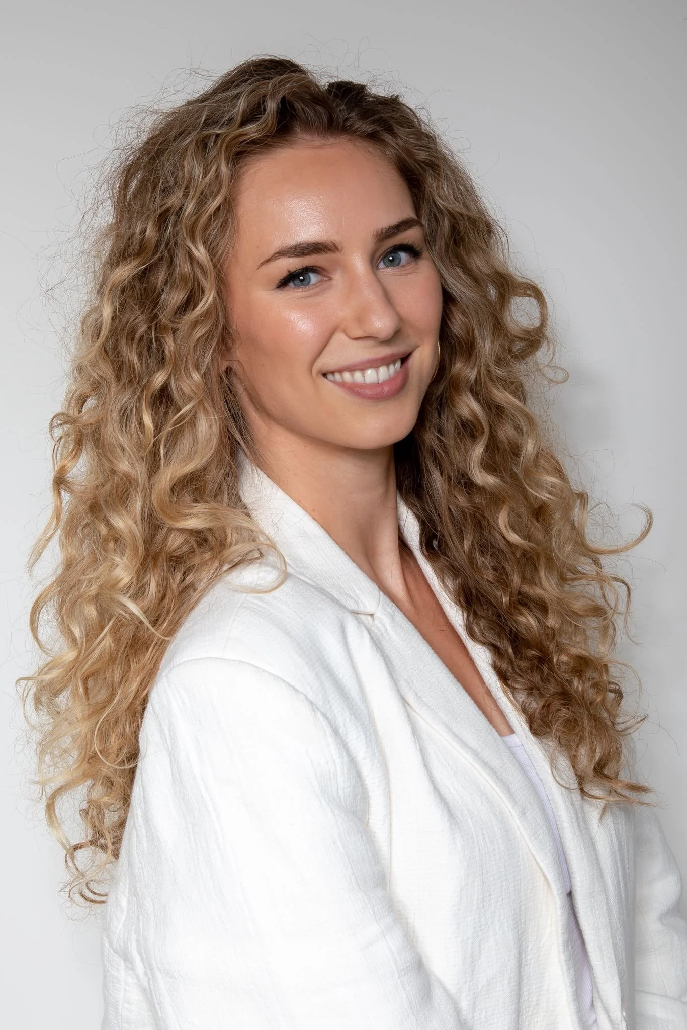 Portrait of a woman with long, curly blonde hair, blue eyes, and light makeup, wearing a white blazer, smiling against a plain gray background.