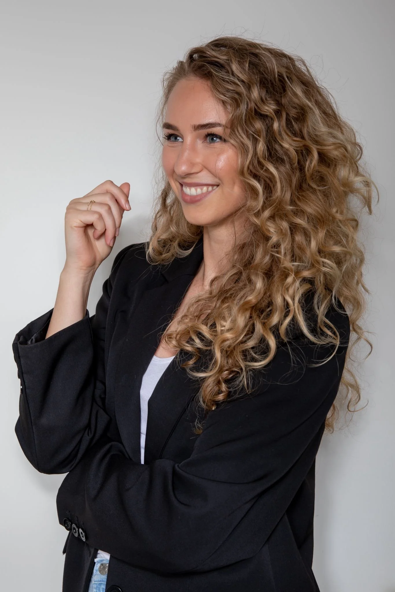 A young woman with long, curly blonde hair smiling and wearing a black blazer over a white top, standing against a plain light-colored background.