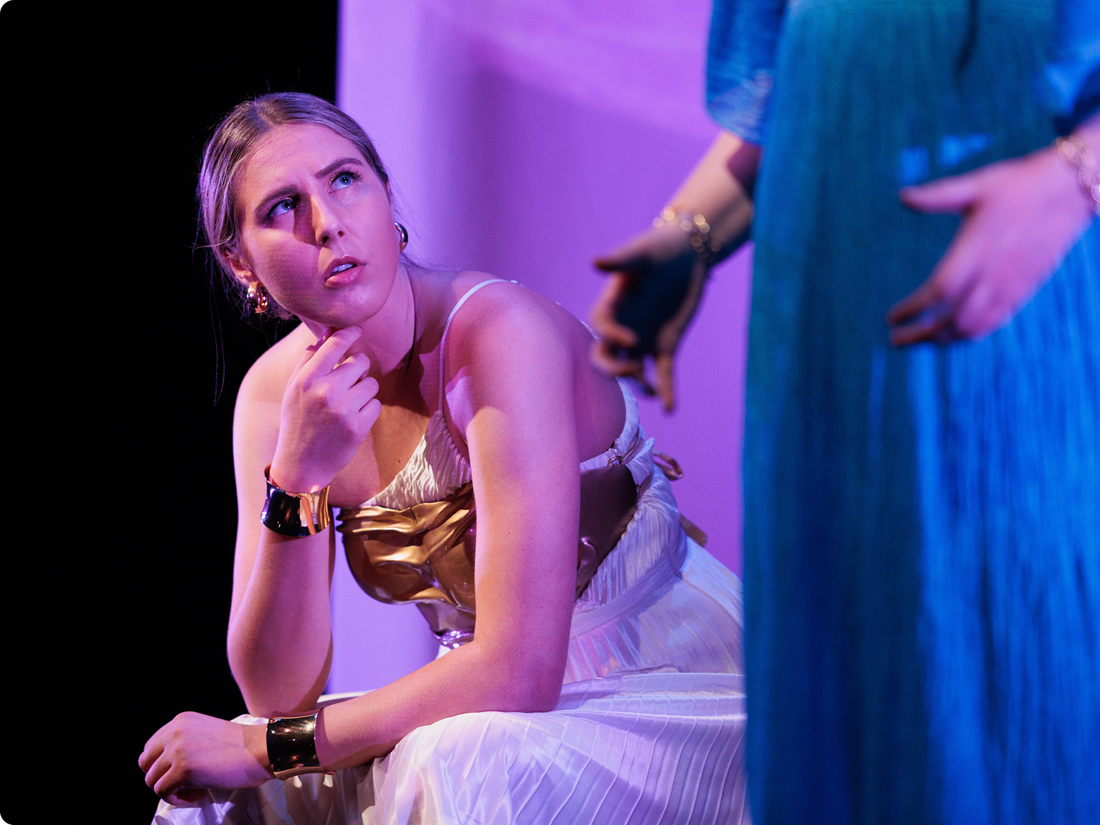 A young woman with blonde hair sitting on a stage, looking up and appearing concerned or contemplative. She is wearing a striped dress with a metallic top and gold accessories, and is gazing at another person standing nearby, who is holding a phone.