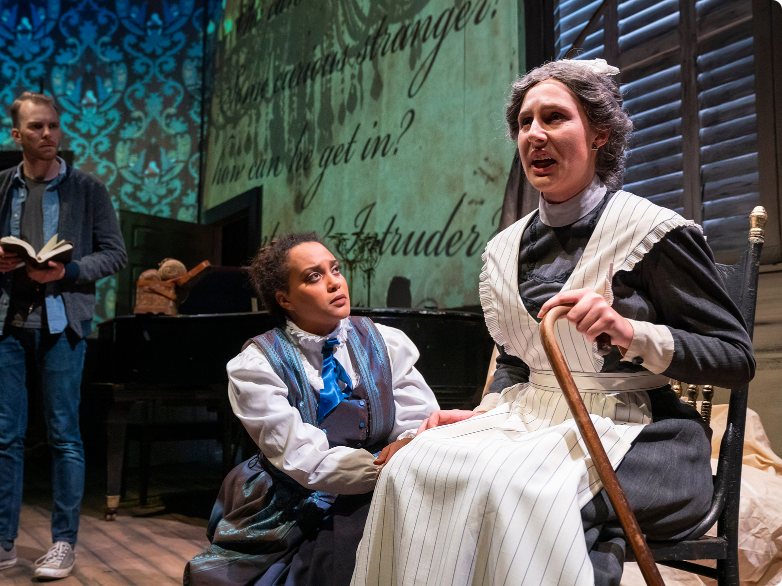 The scene shows three actors performing in a theatrical play set in a historical period. The woman on the right, dressed as a kind older woman with gray curly hair, is sitting on a chair, holding a cane, and appears to be speaking passionately. The y