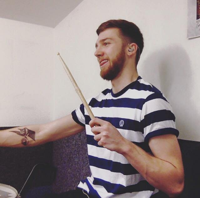 A young man with a beard and short hair, wearing a striped t-shirt, holding a drumstick in his right hand, sitting in a room with white walls.