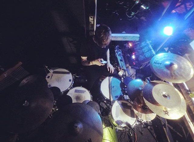 A drummer playing an electronic drum kit on stage with colorful stage lighting.
