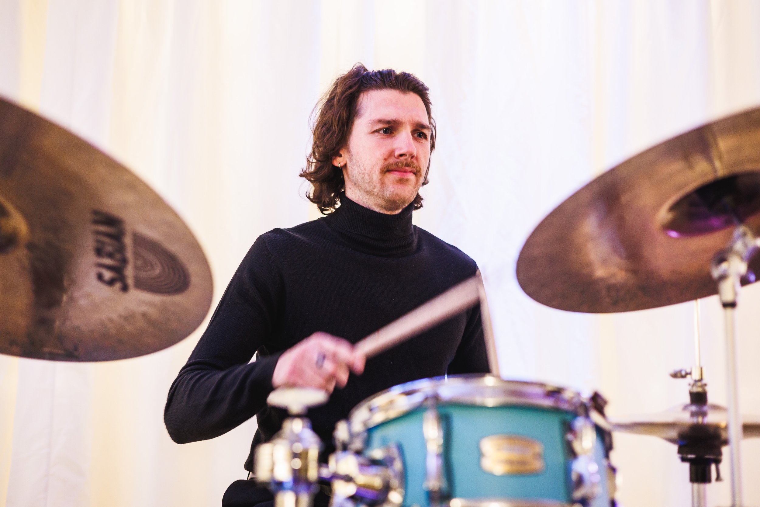 A man with wavy brown hair and a mustache, wearing a black turtleneck, playing a teal drum set with cymbals.