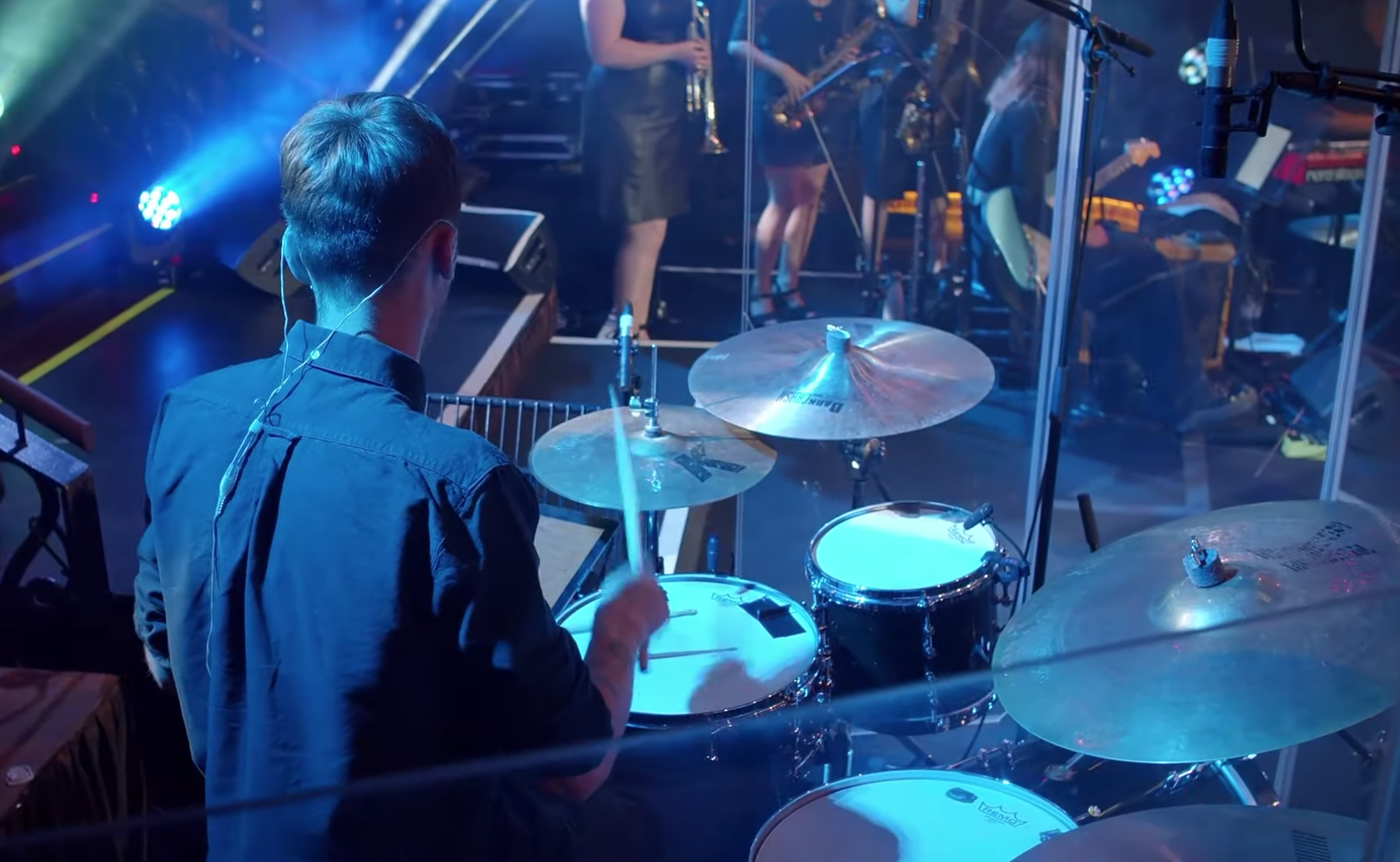 Back view of a drummer playing a drum set on stage during a live concert, with band members playing instruments behind a transparent divider and stage lights illuminating the scene.