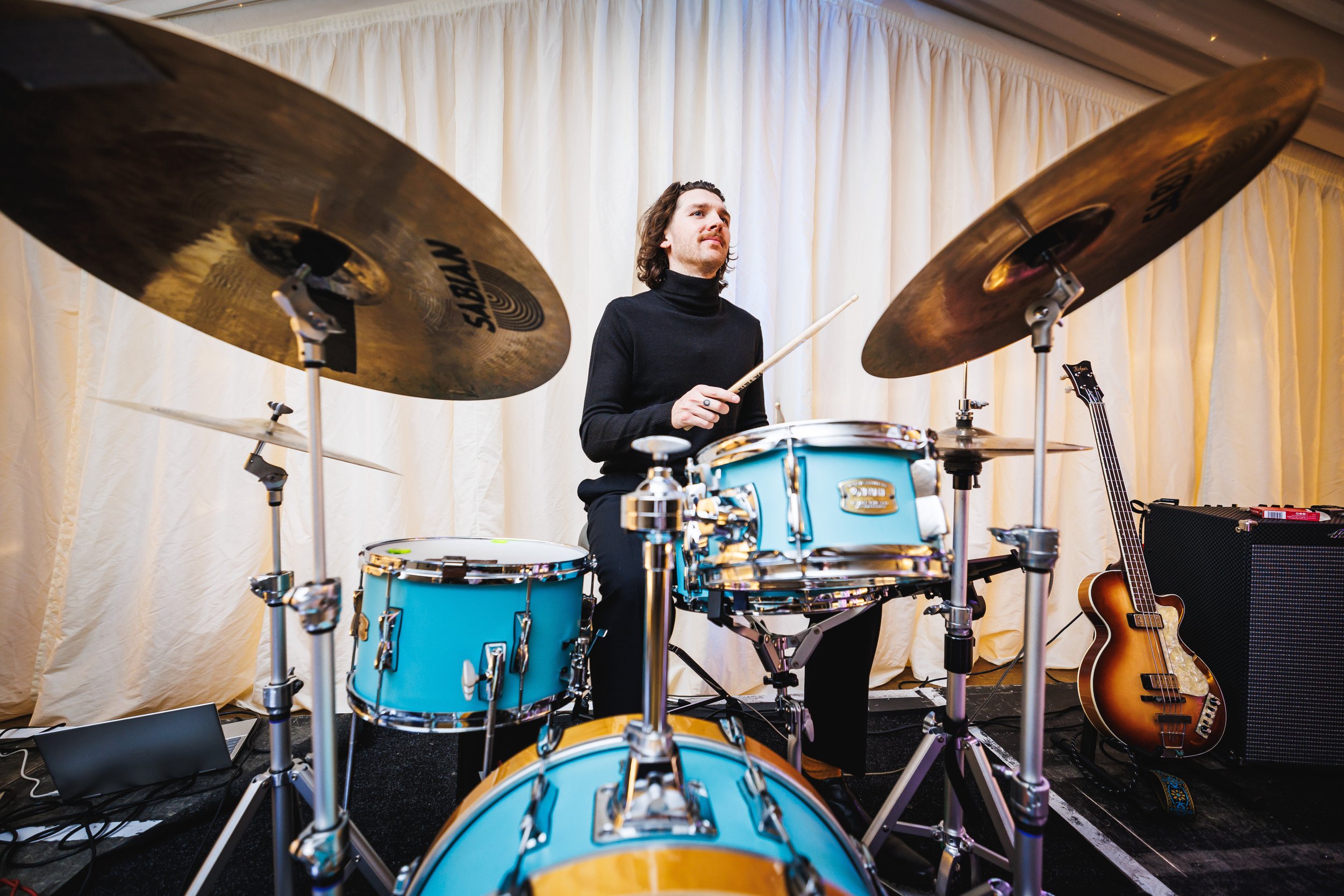 A man playing a blue drum set with cymbals, holding a drumstick, in front of beige curtains, with a guitar and amplifier nearby.