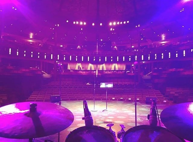 Empty stage with microphones and equipment, surrounded by an auditorium with rows of seats, under purple stage lighting.