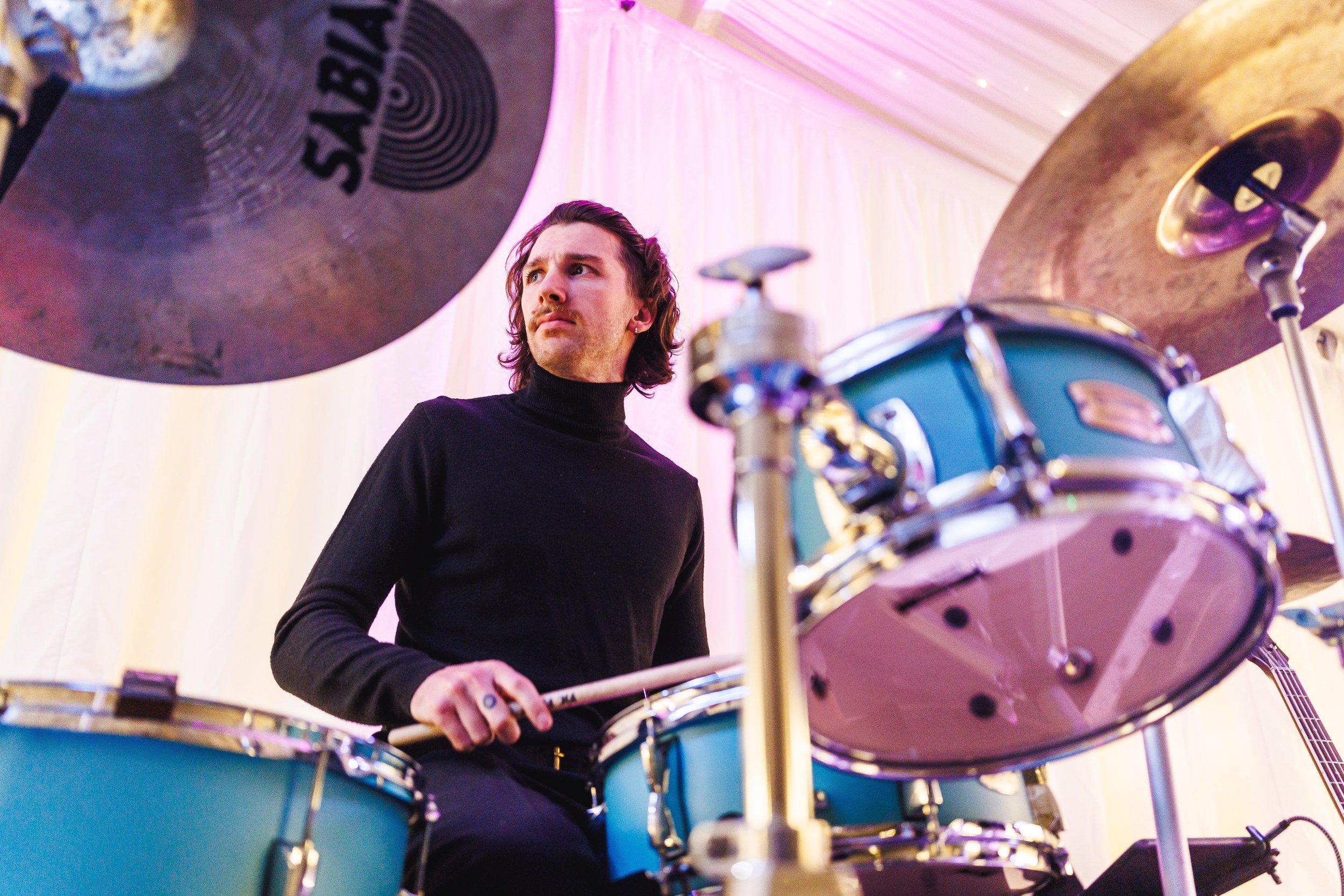 A man with wavy brown hair and a mustache, wearing a black turtleneck, is sitting behind a light blue drum set, holding a drumstick in his right hand. He appears to be looking intently, possibly at something in front or above him. The background has
