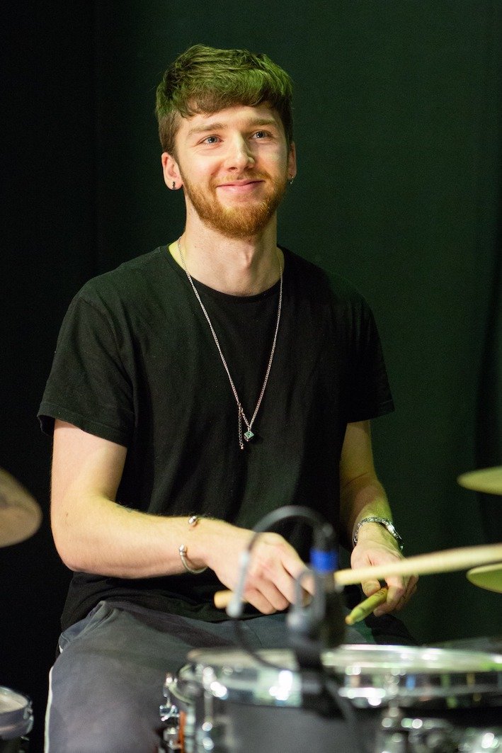 A young man with light brown hair and a beard smiling while sitting behind a drum set, holding drumsticks, in a dark room.