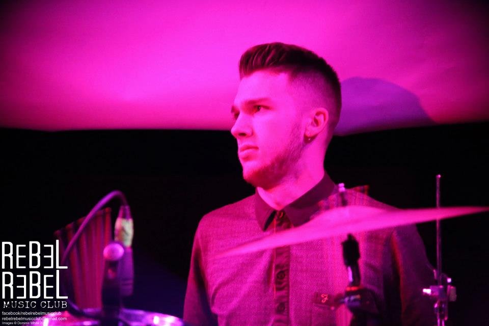 A young man with short hair and a beard plays drums in a music club, illuminated by pink and purple lights.