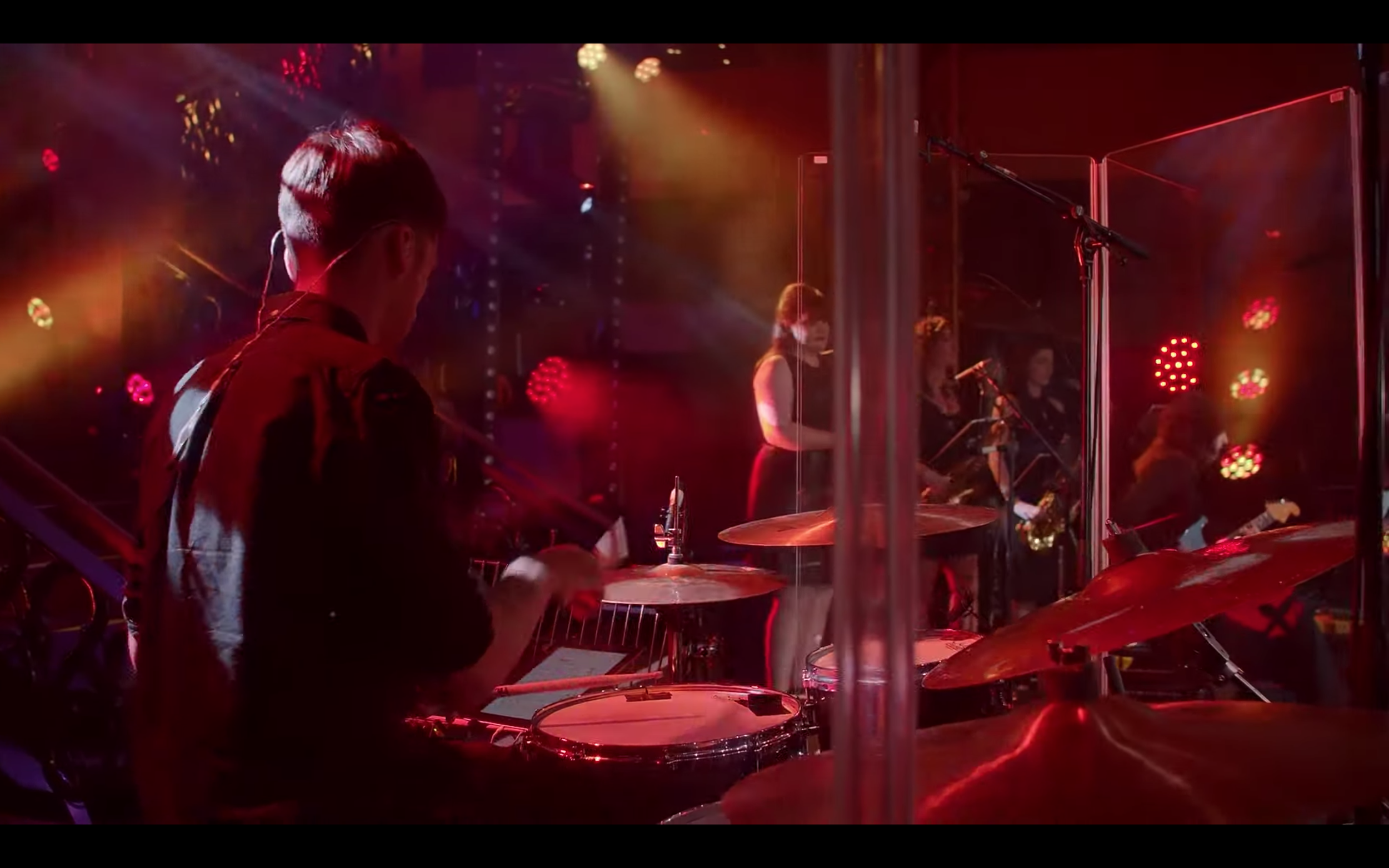 Musicians performing on stage with drums, a vocalist, and instrumentalists under colorful stage lighting.