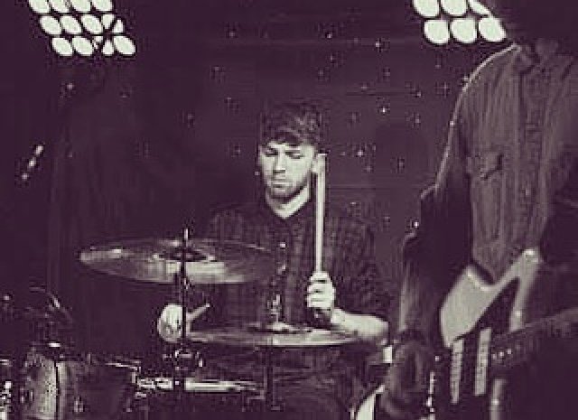 Young man playing drums on stage with a person playing guitar partially visible on the right, stage lights overhead.