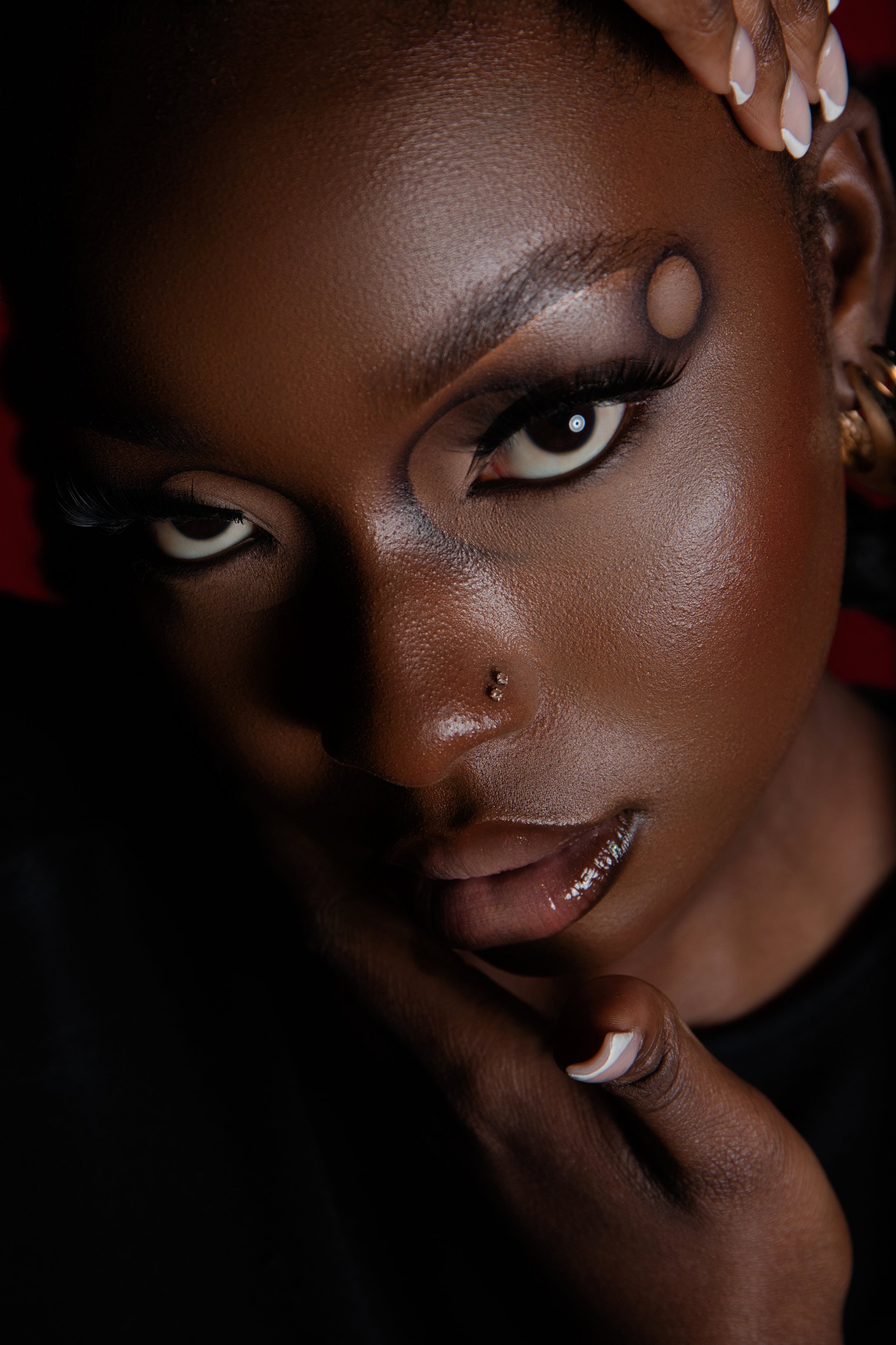 Close-up of a woman with dark skin, bold makeup, and jewelry, resting her face on her fingers.