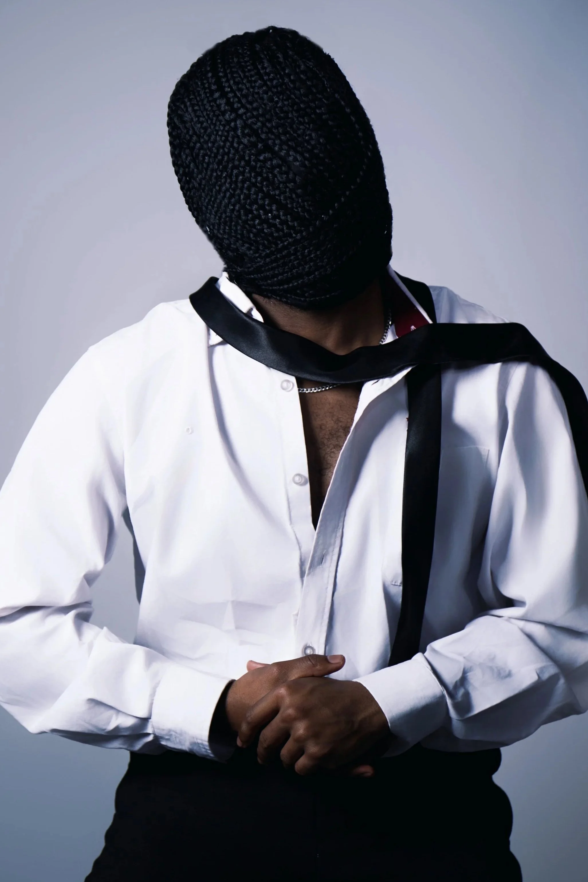 Person with braids wearing a white shirt with an open collar and a black scarf, posing against a plain background.