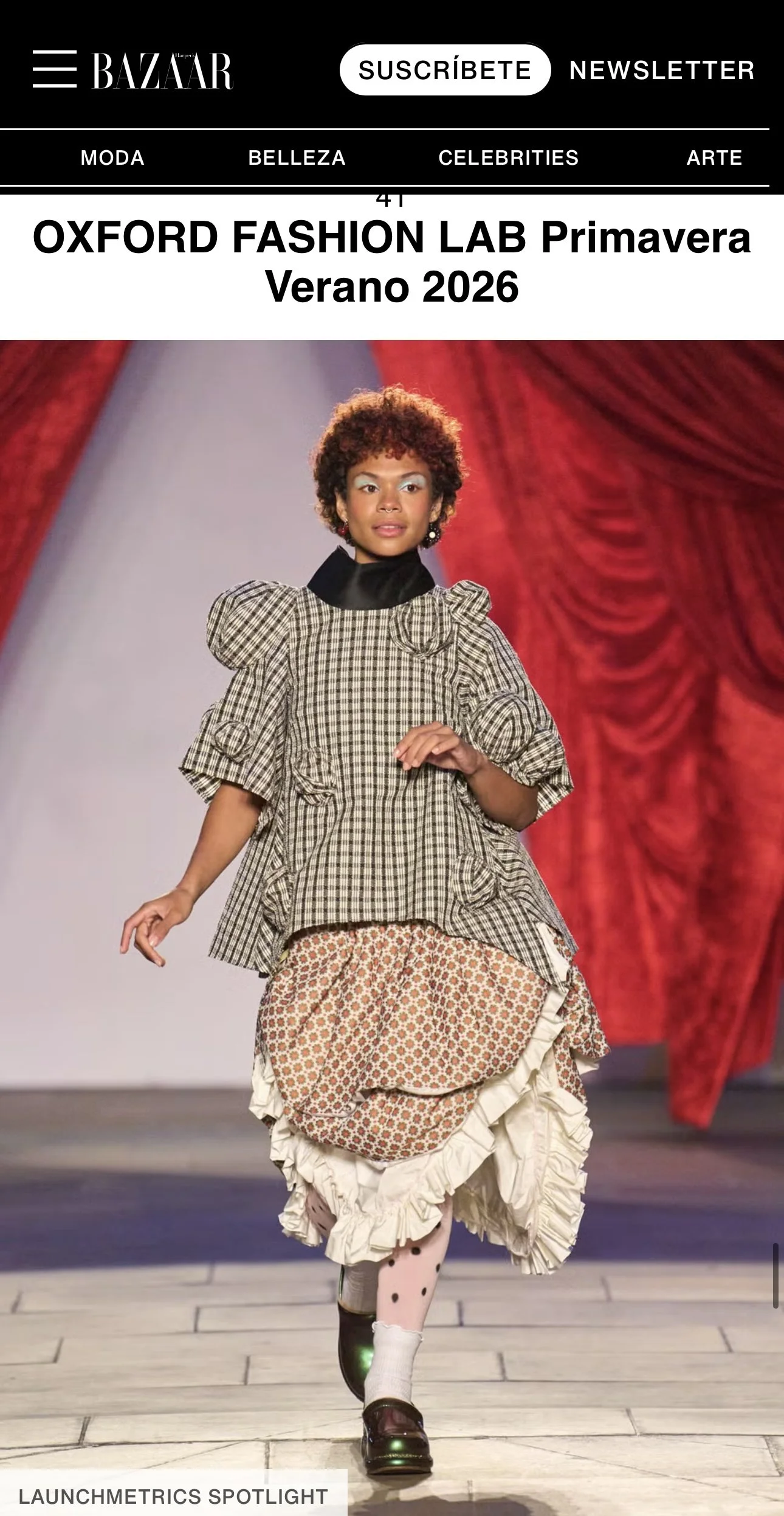 A model walking down a runway wearing a mixed-pattern outfit at the Oxford Fashion Lab Spring summer 2026 show, with a red curtain background.