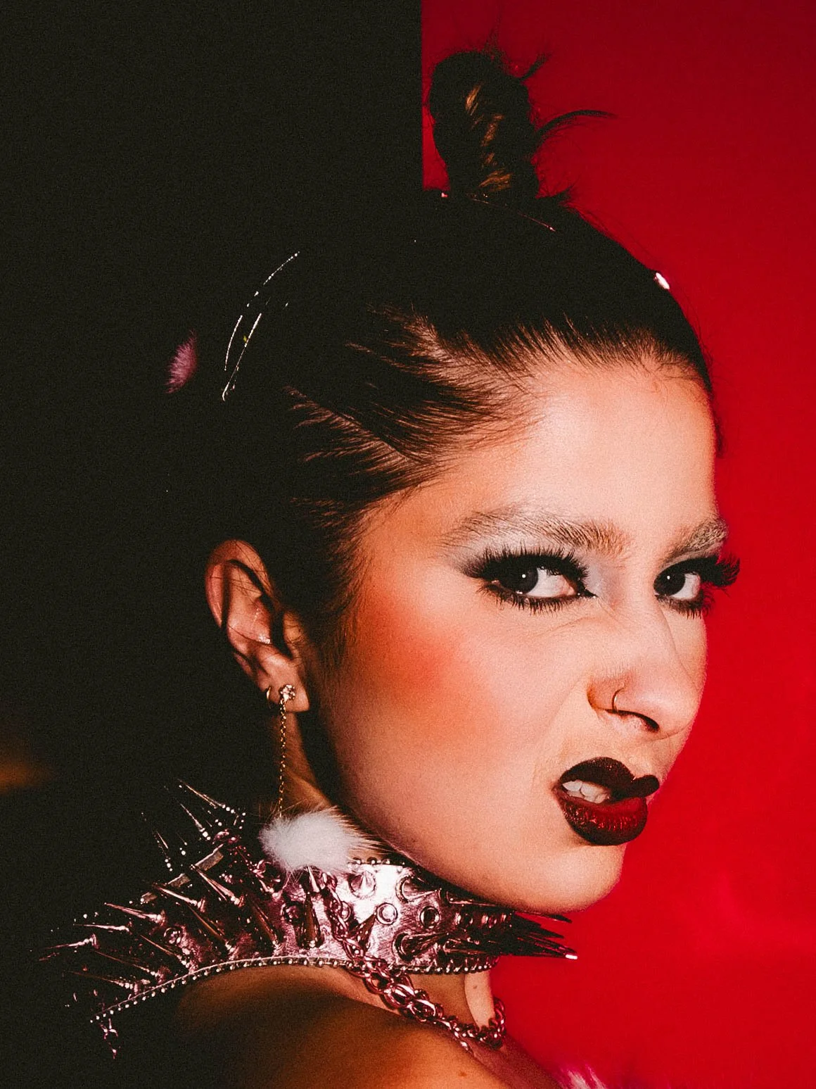 A woman with dark hair styled in a high bun, wearing dark eye makeup, black lipstick, multiple earrings, and a metallic spiked collar, against a red background.