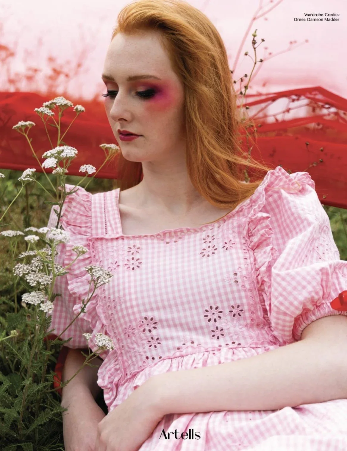 A young woman with red hair and fair skin, wearing a pink gingham dress with eyelet cutouts, sitting outdoors among white wildflowers, with a red umbrella behind her.