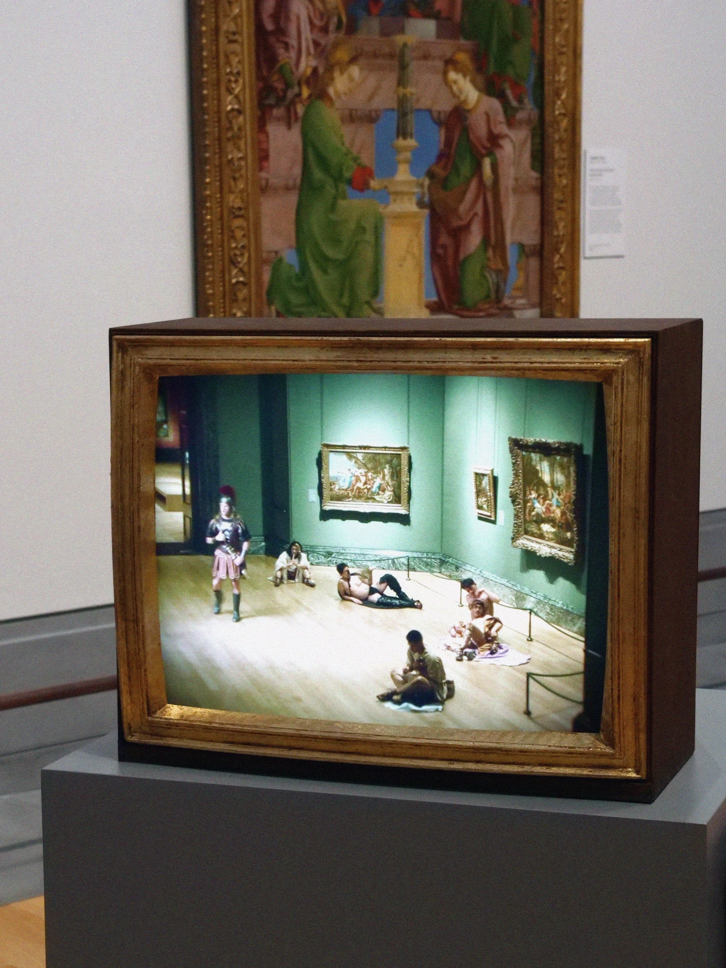 A museum exhibit showcasing a diorama of a daytime scene in an art gallery where children and adults are sitting on the floor, observing artwork and a performer dressed in a costume resembling a museum guard.