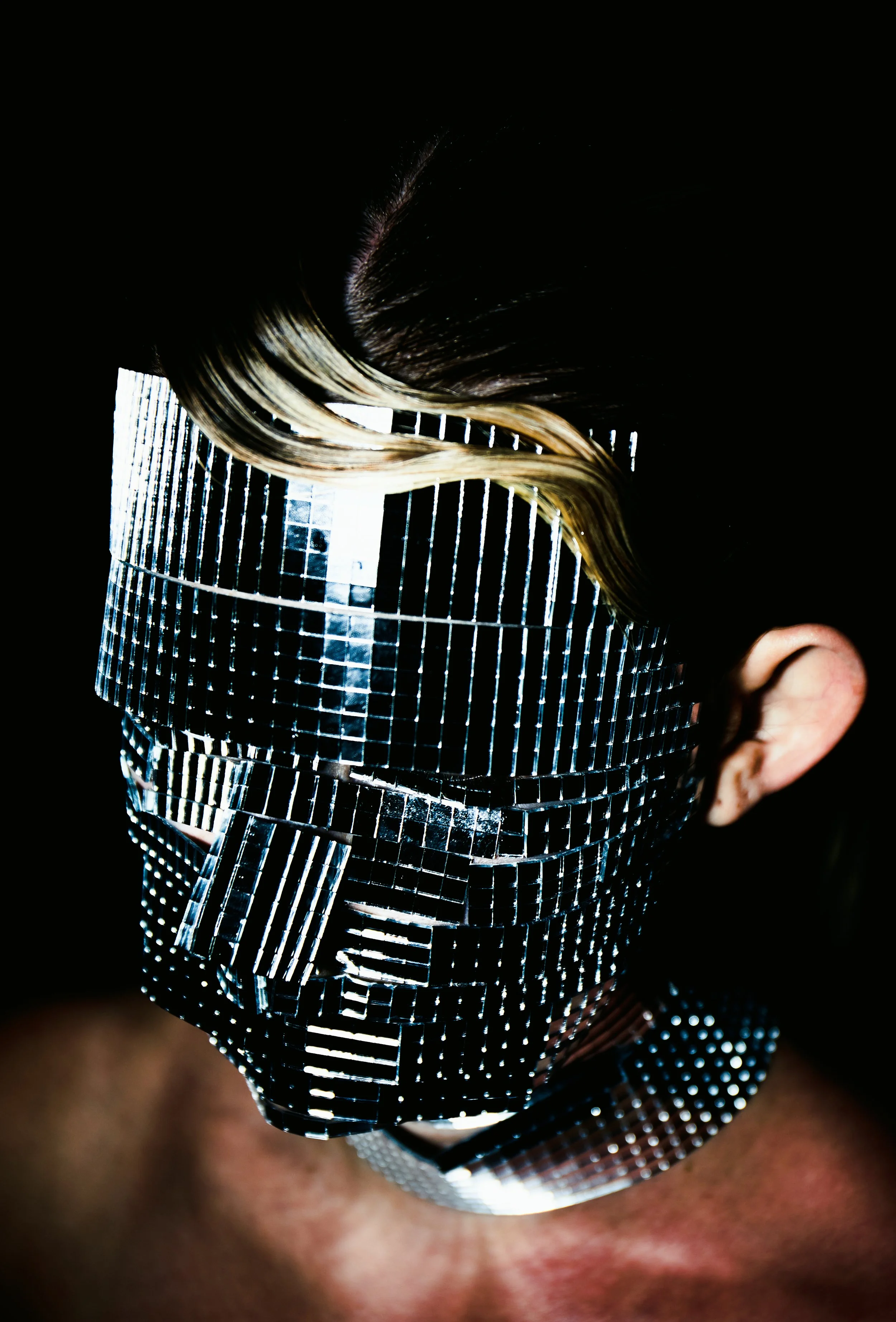 Person wearing a face mask covered with small reflective mirror tiles, with highlighted hair, against a black background.