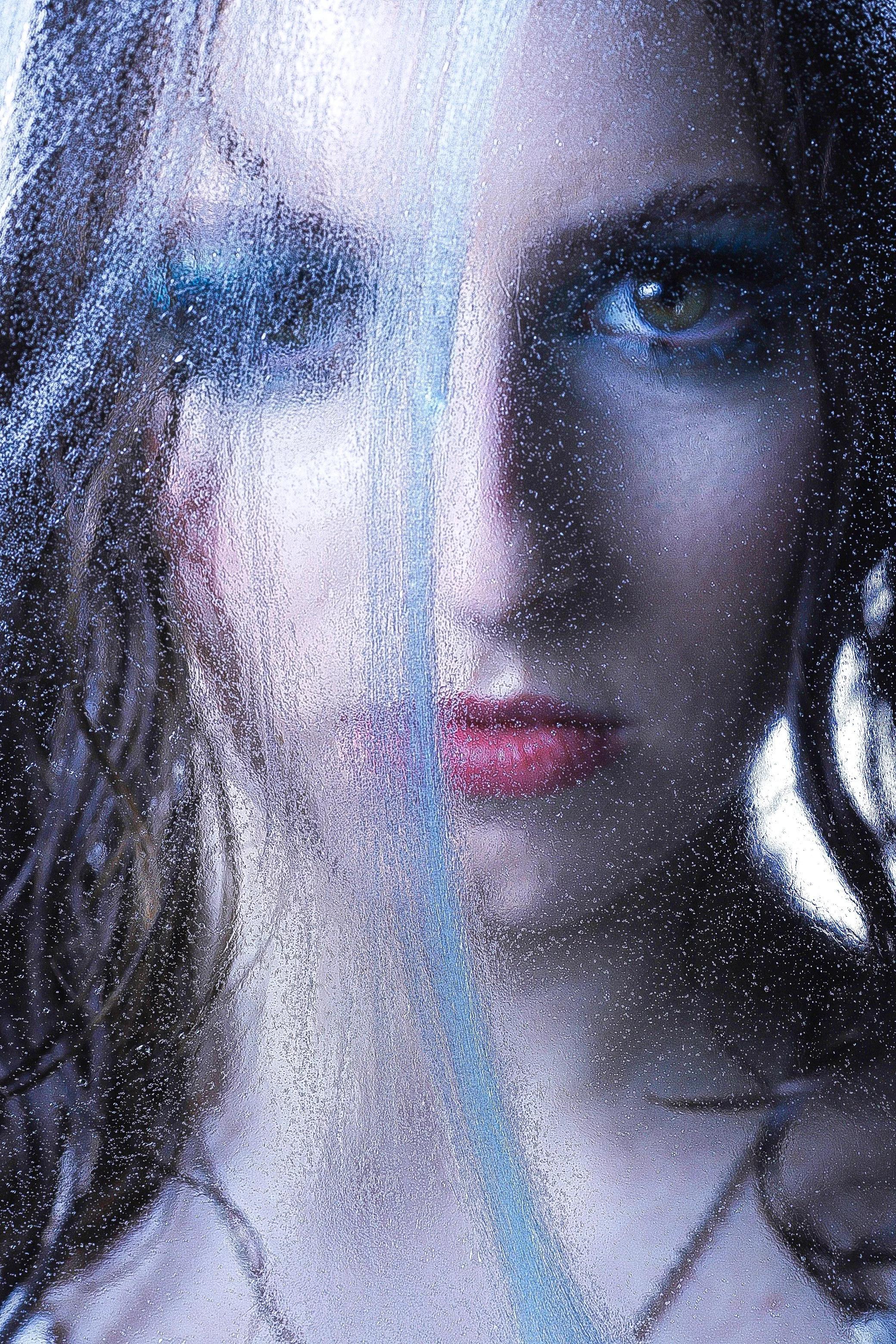 Close-up of a woman's face seen through a rain-covered glass window, with her left eye partially visible and a piercing gaze.