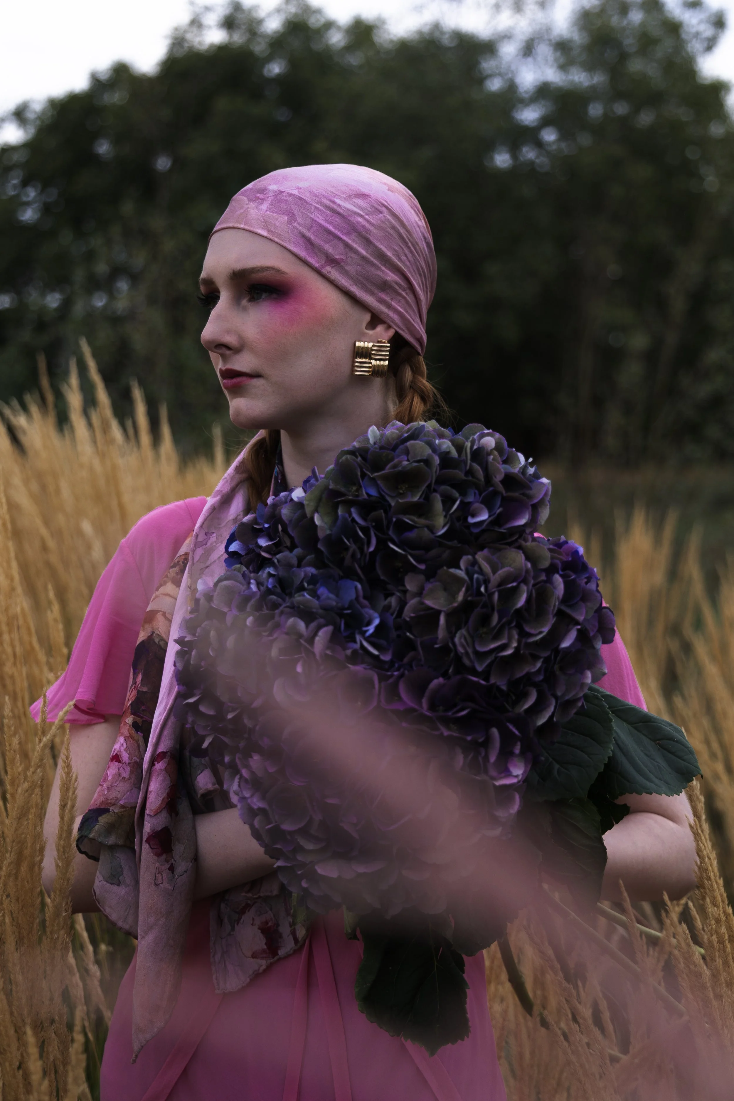 A woman with red hair wearing a pink headscarf and pink dress, holding a large purple hydrangea flower, standing in a field of tall, golden grass with green trees in the background.