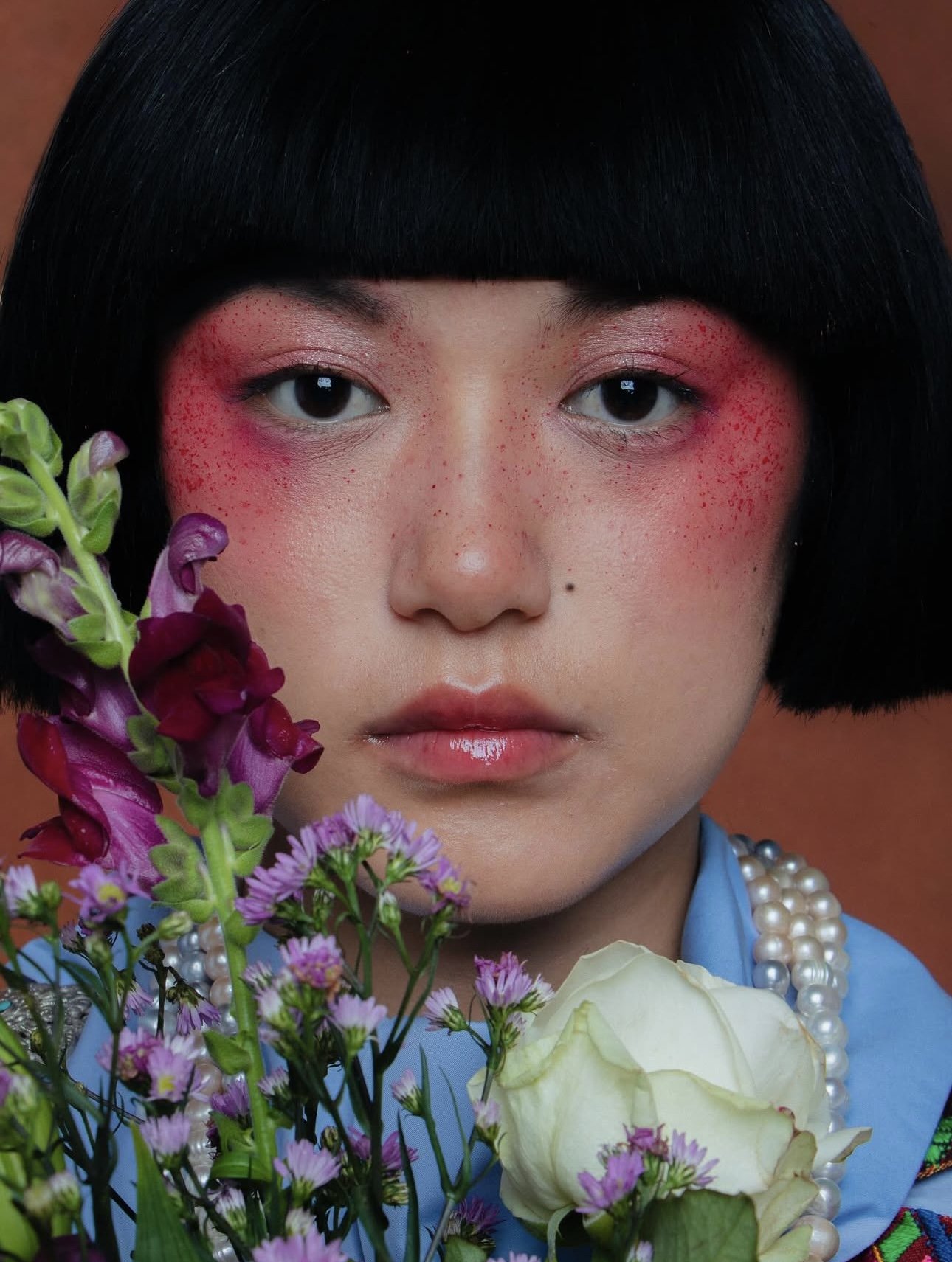 A close-up portrait of a woman with short black hair and makeup featuring red eyeshadow and freckles, holding colorful flowers including purple, white, and pink blossoms, with a pearl necklace and a blue top.