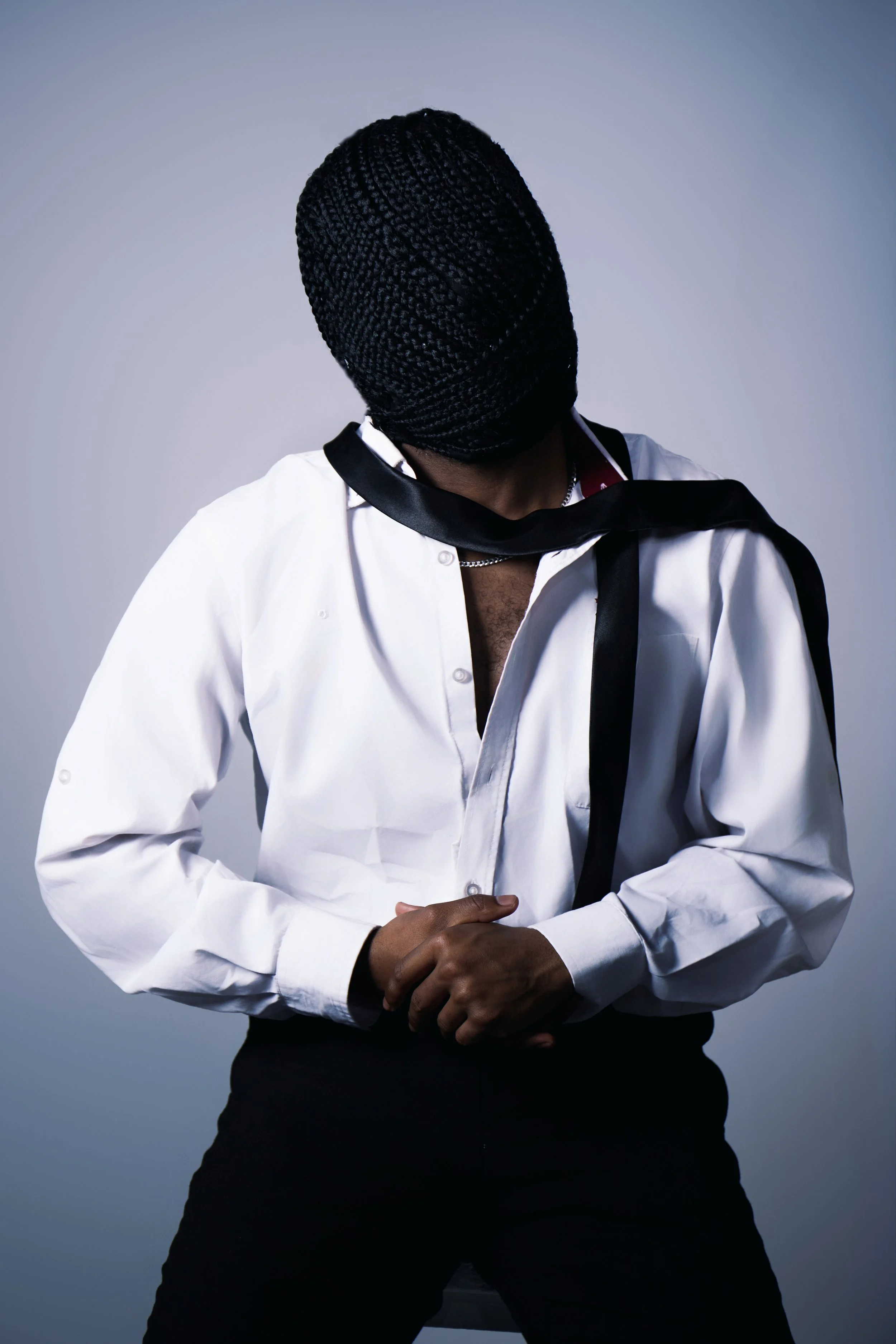Person with braids wearing a white shirt with an open collar and a black scarf, posing against a plain background.