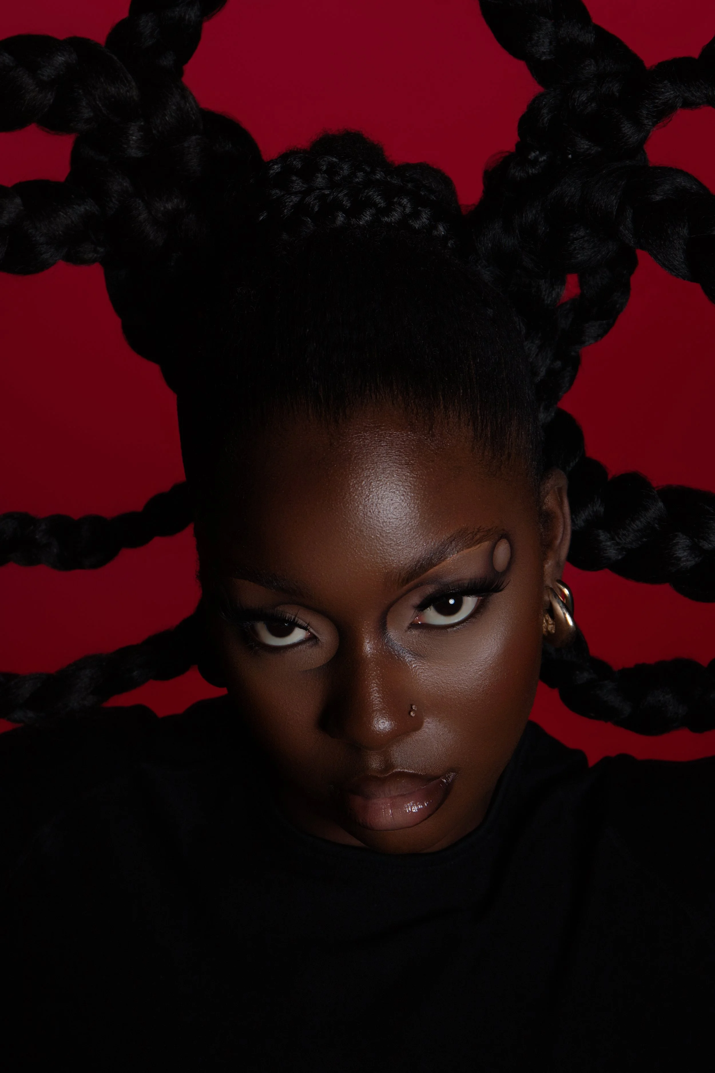 Close-up portrait of a black woman with dark skin and braided hair. She has intense eye contact, makeup, a nose piercing, and glossy lips. She is holding her head with both hands against a red background.