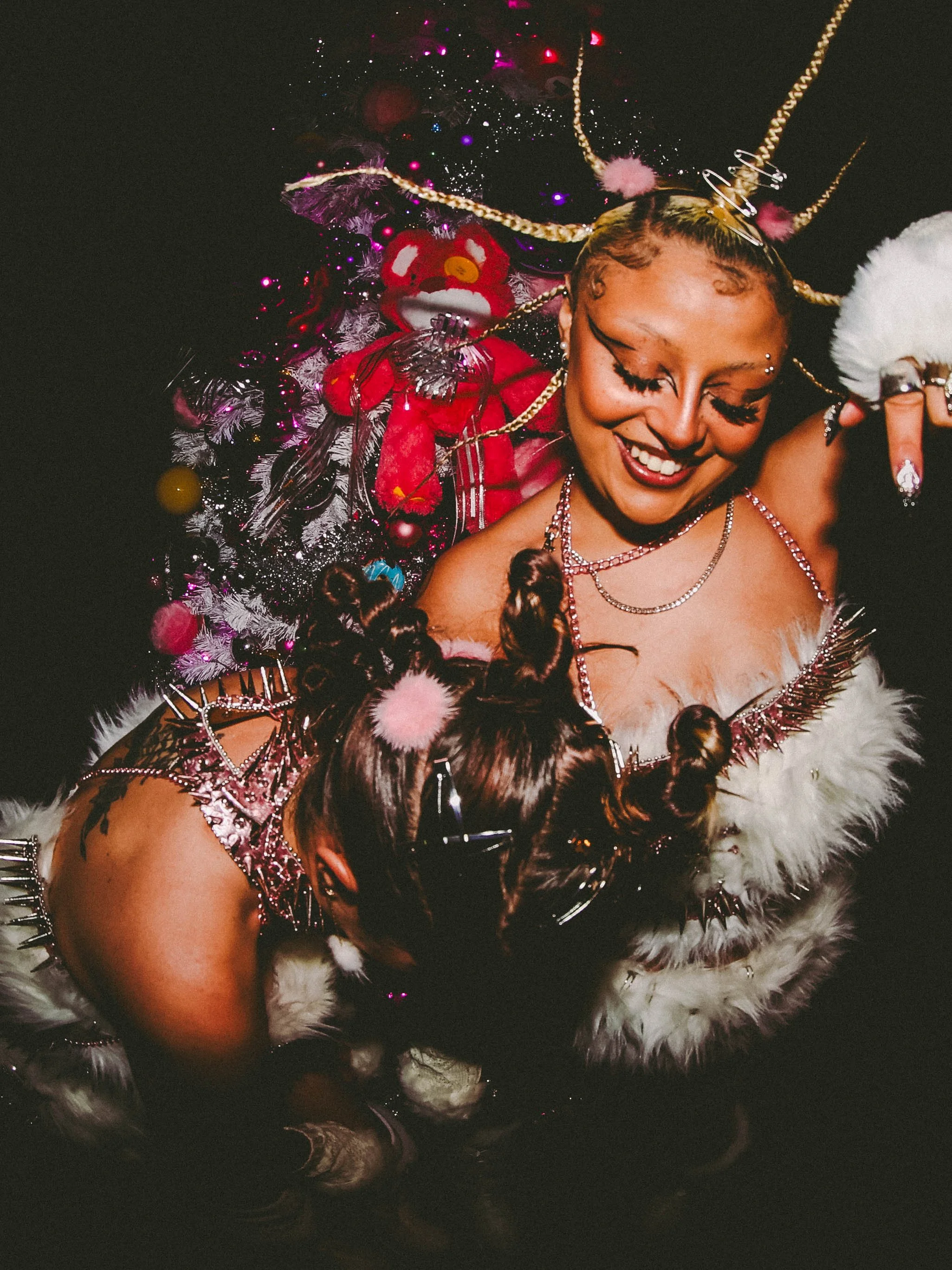 A person dressed in festive, glittery clothing with jewelry and makeup, smiling and leaning towards another person with styled hair and holding a phone, standing in front of a decorated Christmas tree with ornaments and a red plush teddy bear.