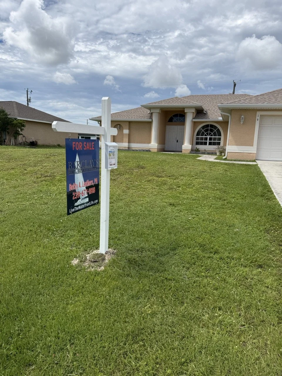 Front yard of a house with a 'For Sale' sign in the grass, and a beige single-story house with a front door, window, and garage visible.