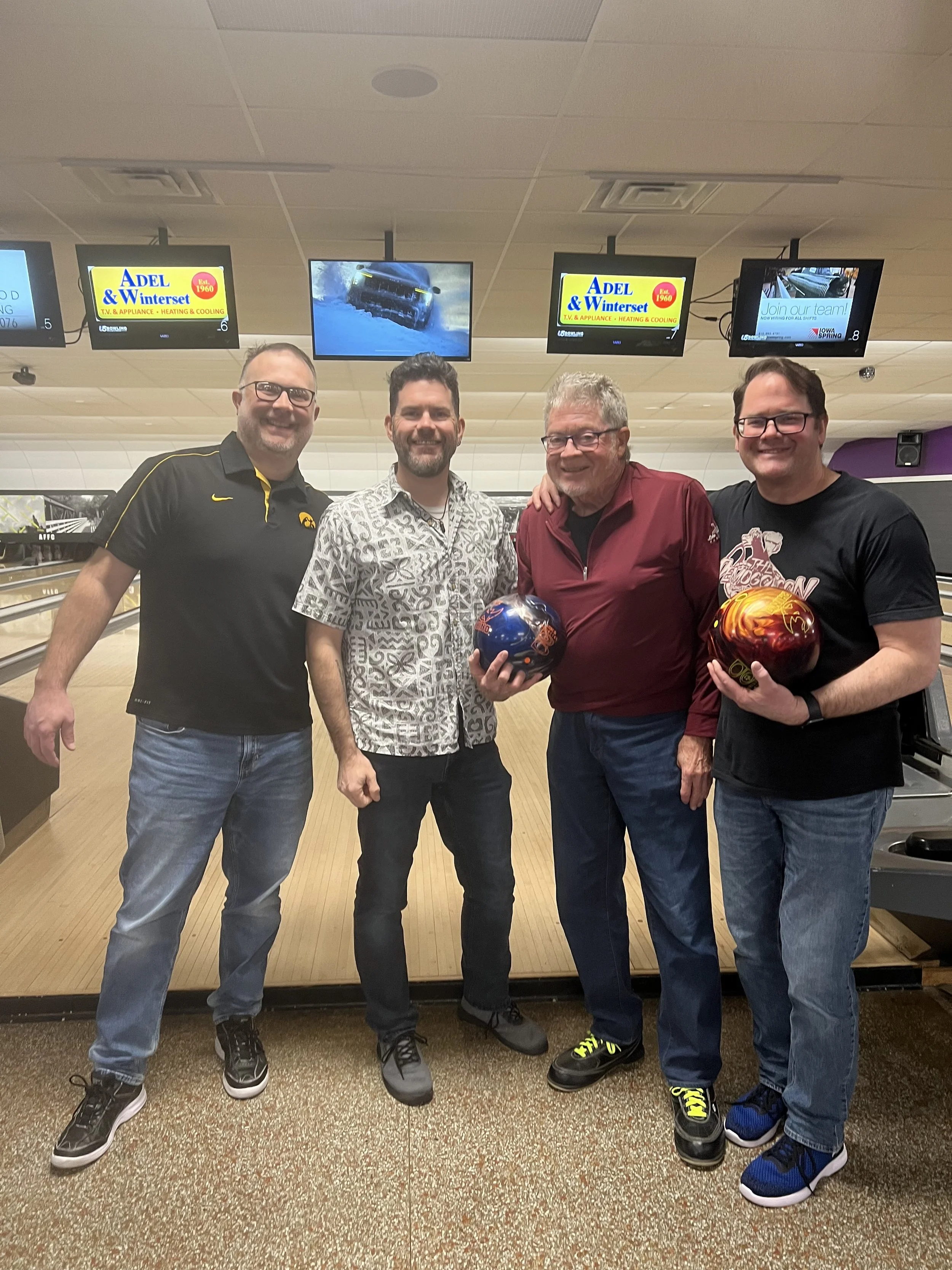 Four men standing together at a bowling alley, each holding a colorful bowling ball, smiling and posing for the photo.