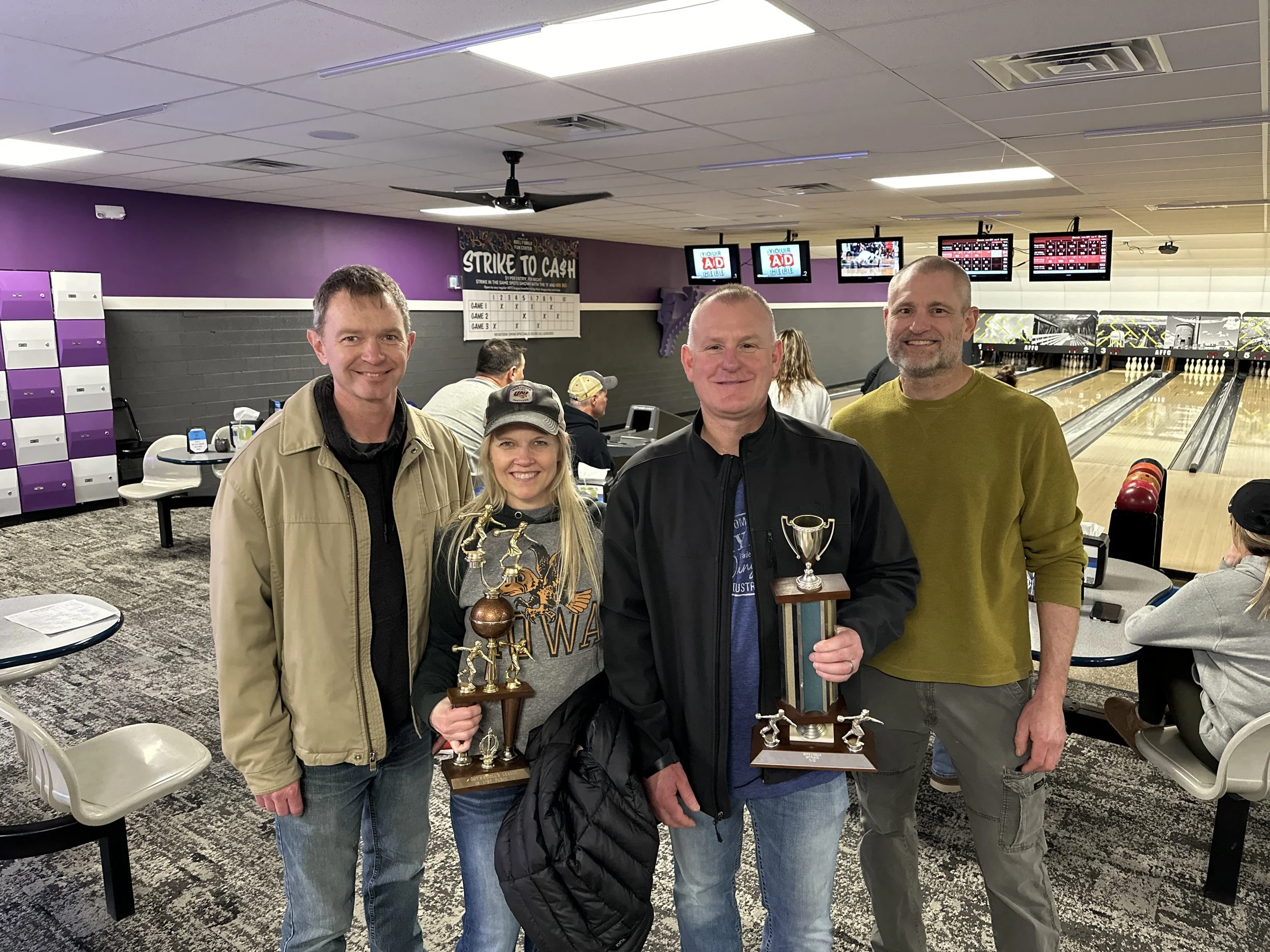Four people standing in a bowling alley, two of them holding trophies. The background shows bowling lanes, other people, and electronic score screens.