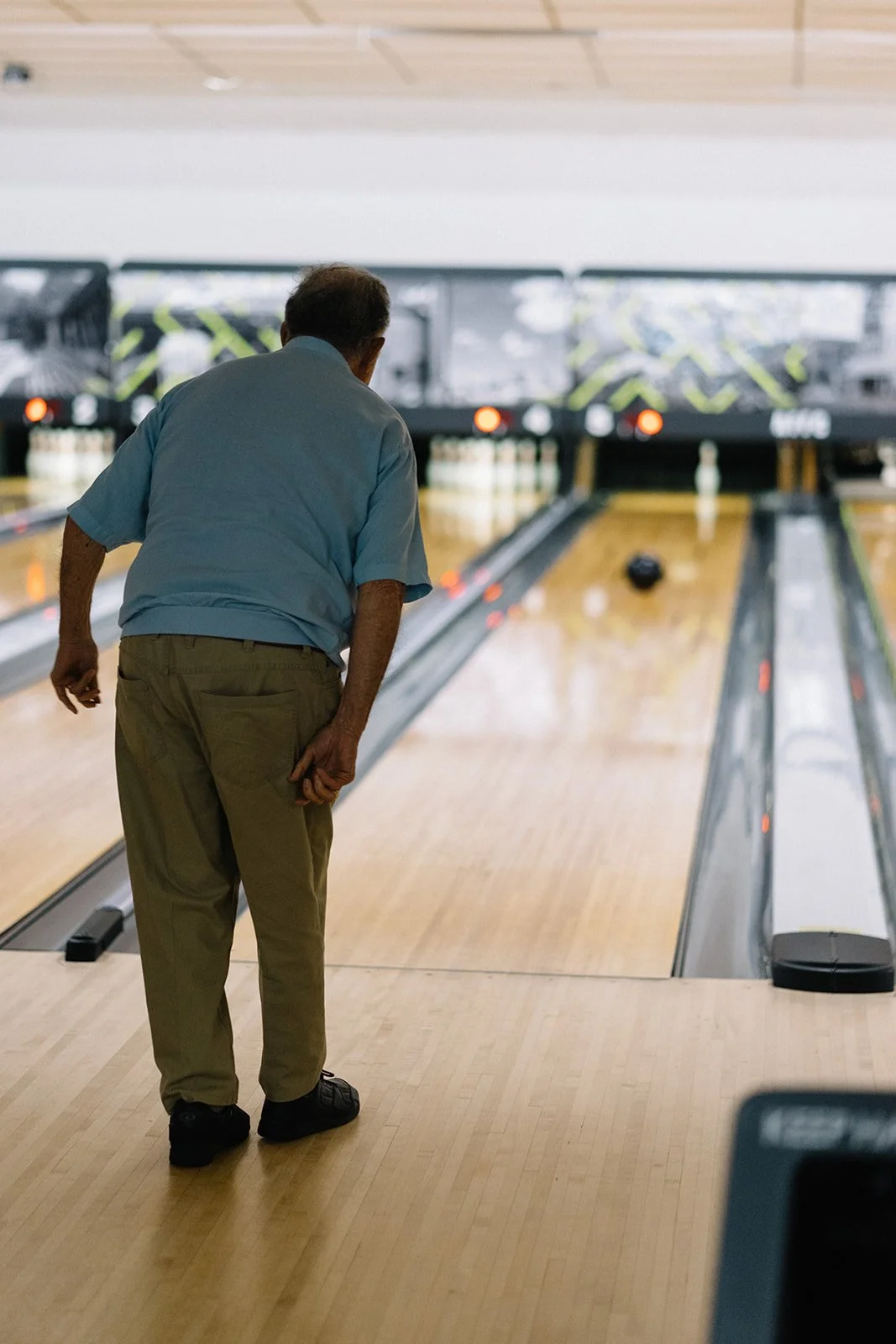 An elderly man in a light blue shirt and khaki pants is bowling on an indoor lane, preparing to throw the ball.