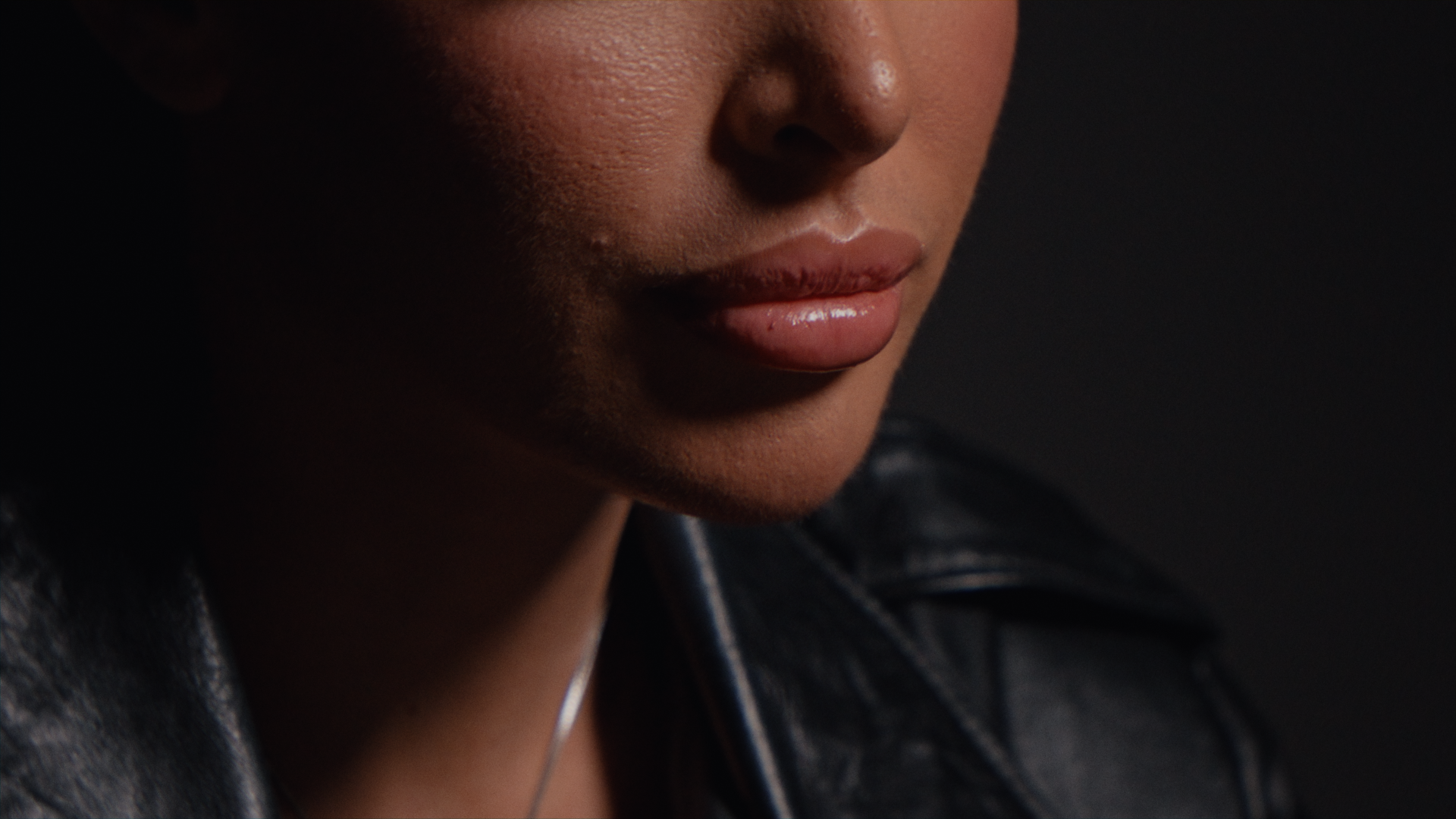 Parte del volto di una donna con labbra piene e pelle scura, vestita in pelle nera, sfondo nero.