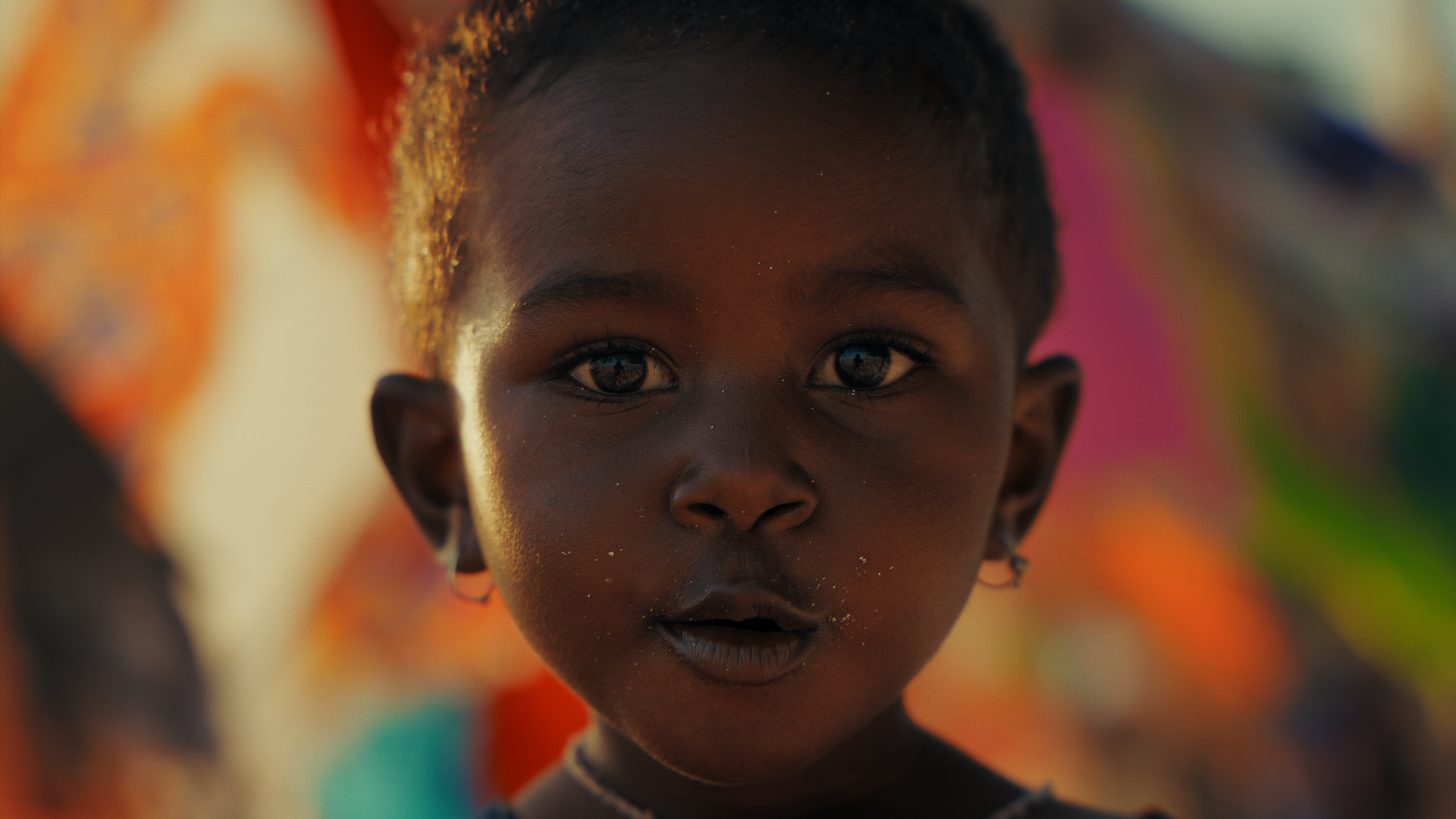 Ragazzo con pelle scura, occhi grandi, capelli corti e orecchini in argento, con sfondo colorato sfocato.