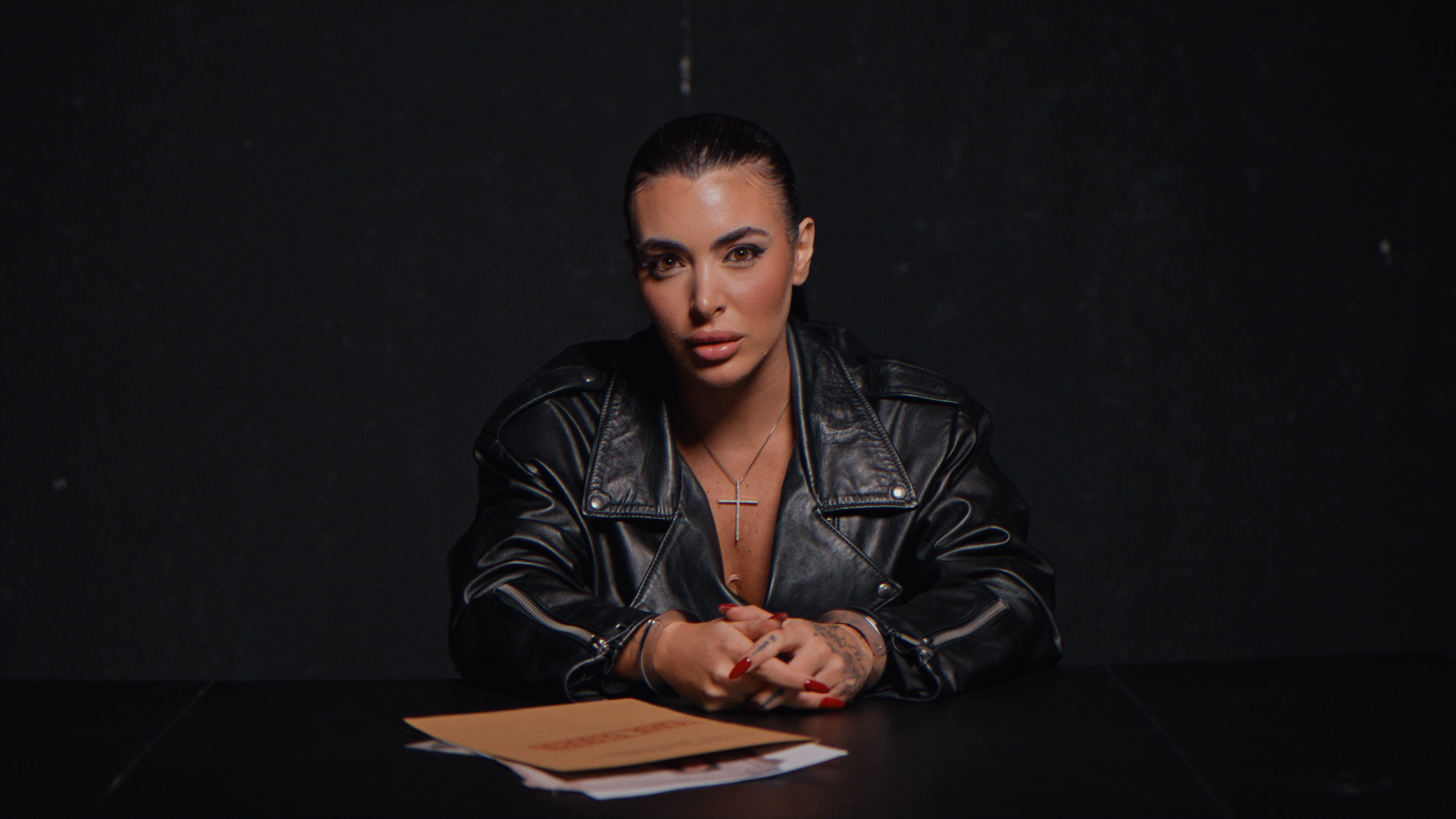 Donna seduta a un tavolo con un vestito nero, mani incrociate e sguardo diretto alla fotocamera, sfondo nero.