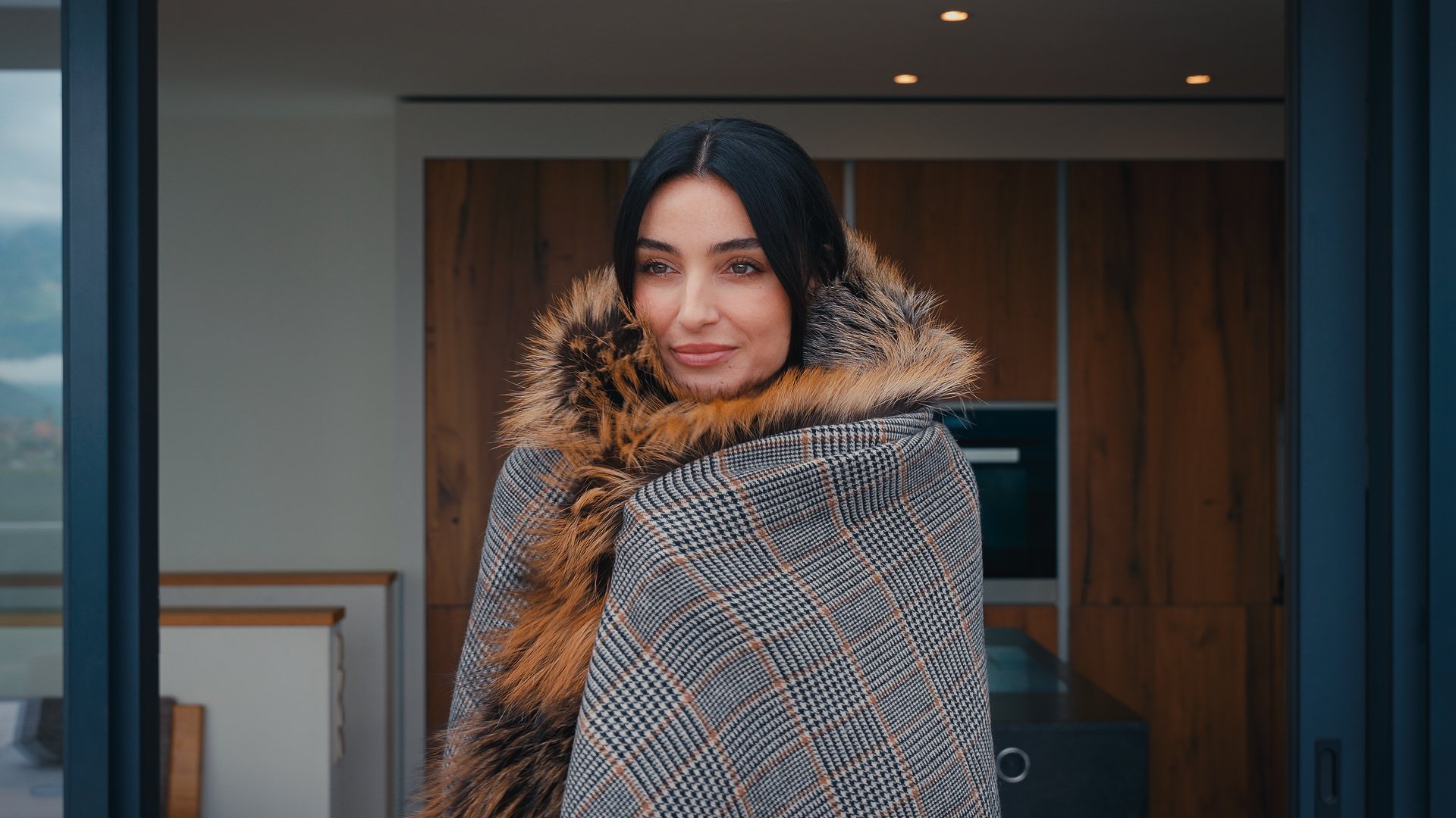 Una donna con lunghi capelli neri, indossa un cappotto con pelliccia intorno al collo e un abito tartan, si trova in un ambiente domestico moderno con mobili in legno.