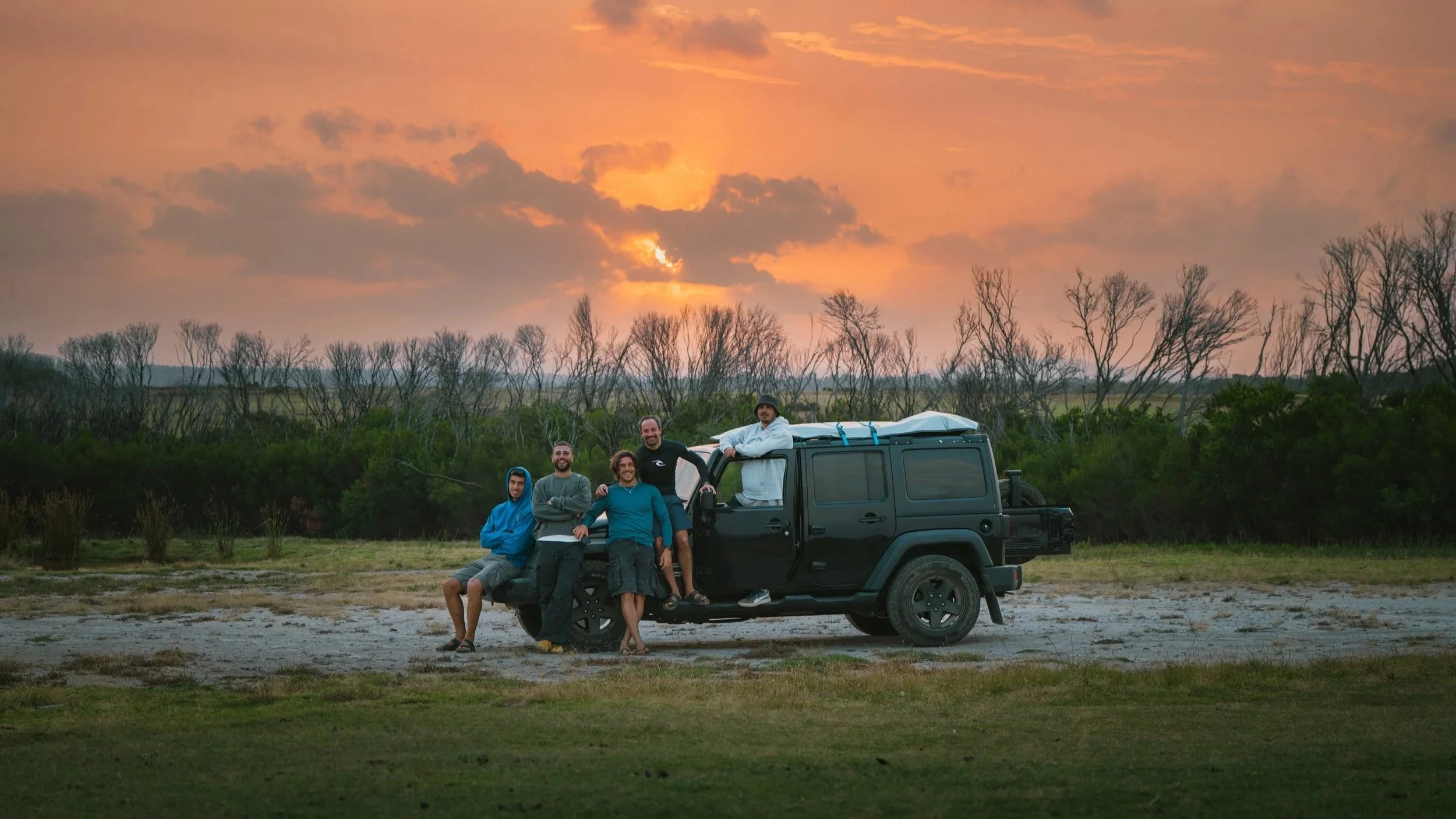 Gruppo di cinque persone che si rilassa e scatta fotografie accanto a un'auto nera in un paesaggio naturale durante un tramonto, con cielo arancione e alberi spogli sullo sfondo.